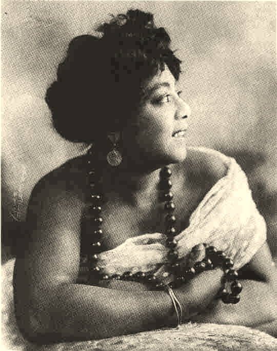 A photograph of American singer Mamie Smith, wearing a lace dress and a large beaded necklace, smiling and looking off-camera.