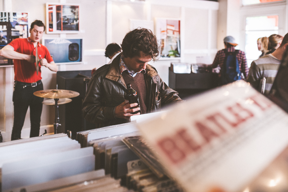 People shopping in a vintage record store. In the exteme foreground, we see a vinyl copy of a Beatles compilation being held up for perusal. In the middleground, a man who appears to be in his twenties, wearing a colroful collared shirt, a V neck sweater and a leather bomber jacket holds a bottled beverage while looking through a rack of vinyl albums. In the background, other shoppers look through separate racks of vinyl albums while a man in a red polo shirt and black jeans assembles a drum kit.