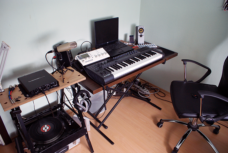 A home recording studio. On the left, a portable workhorse holds a record player, an audio interface, and a microphone on a tabletop tripod, with a pair of over-the-ear headphones hanging on one of the workhorse's vice cranks. In the center of the photo, a collapsble keyboard stand holds a controller keyboard, an outboard synthesizer, a laptop computer and a set of small powered speakers. An empty office chair sits to the right.