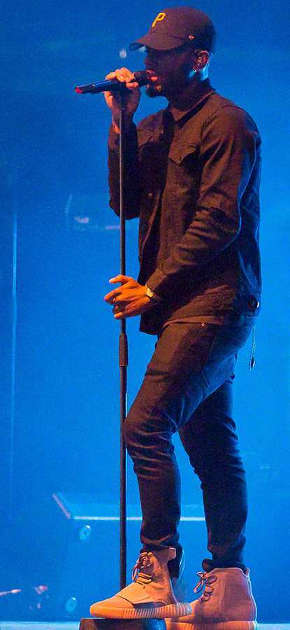Bryson Tiller, wearing a black cap, jacket and jeans and white hightop sneakers, performs on stage holding a mic on a mic stand.