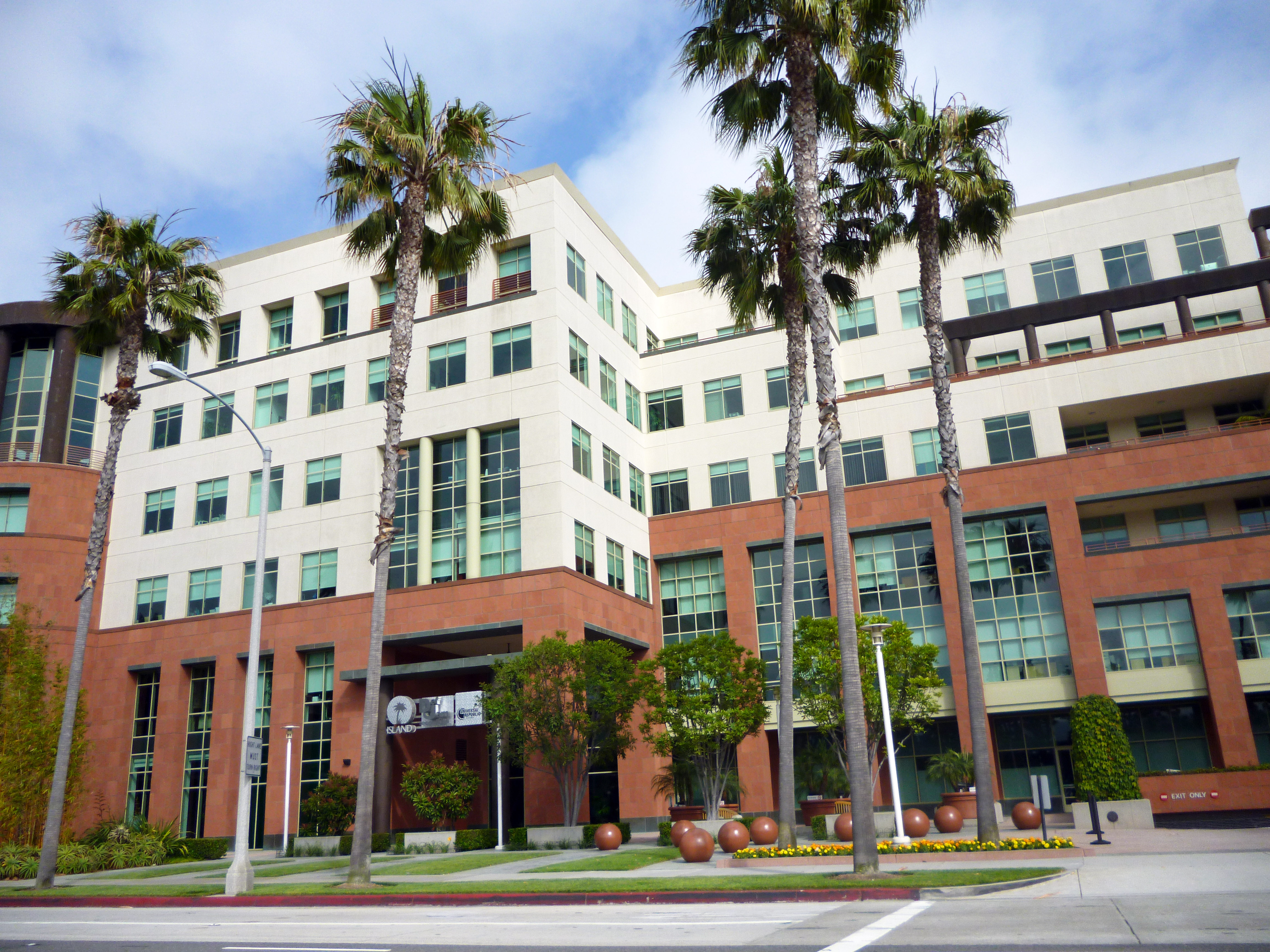 The front of an eight to ten story building. The lower floors have a orange-brown stone facade; the upper floors are white. All levels have rows windows, many with shades partially pulled. The grassy area between the building and the street has five tall palm trees and several shorter trees.