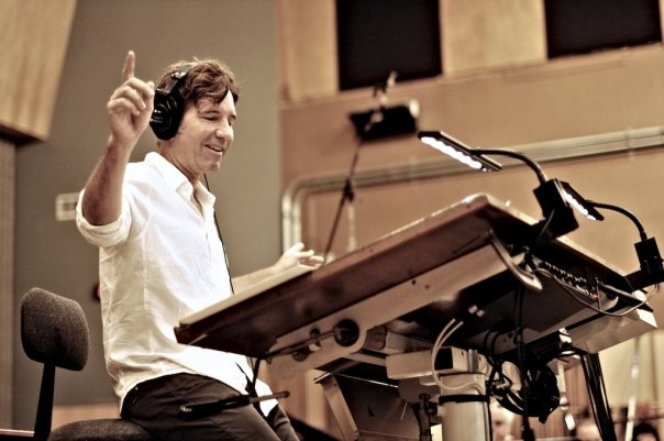 A conductor sits on the edge of a stool with a large music stand in front of him. He is wearing over-the-ear headphones and is smiling while conducting, with both arms held out.