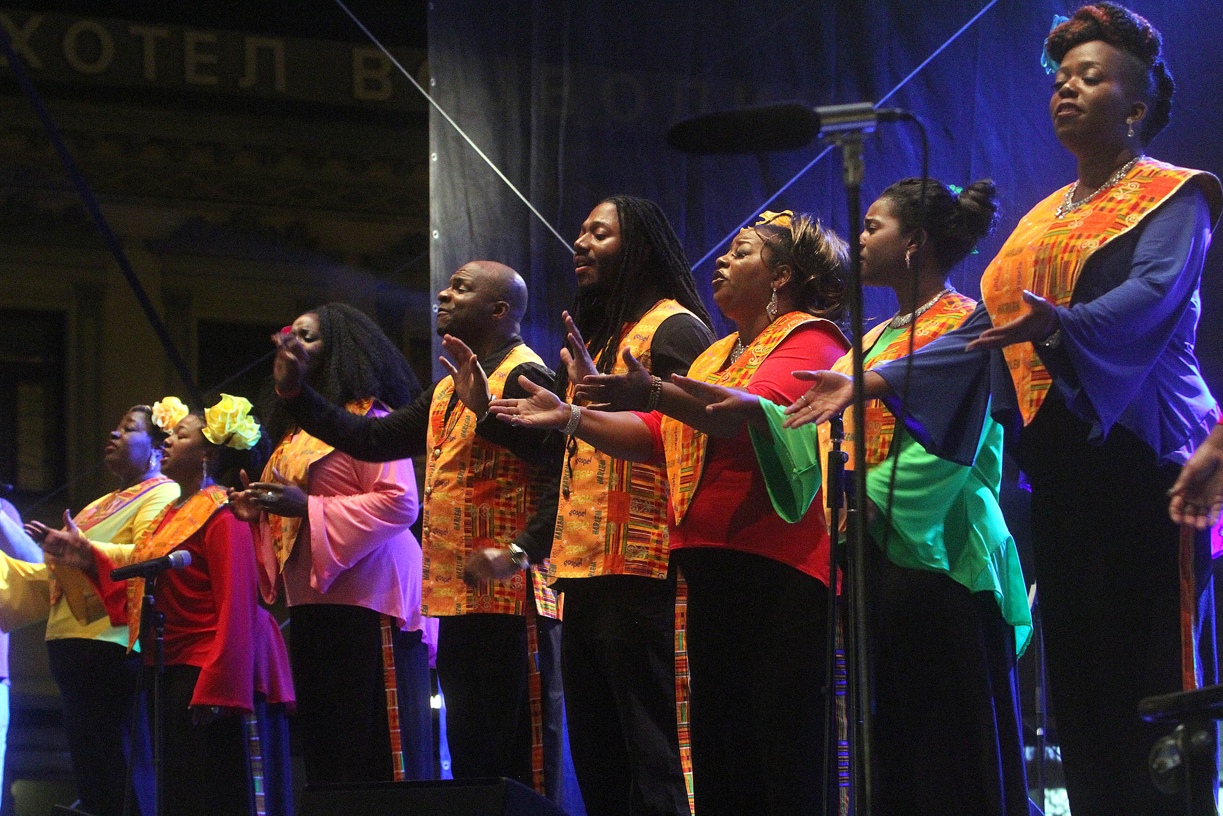 A photograph of eight members of the Harlem Gospel Choir singing on stage, wearing brightly-colored shirts and vests and motioning with their hands as part of the performance.