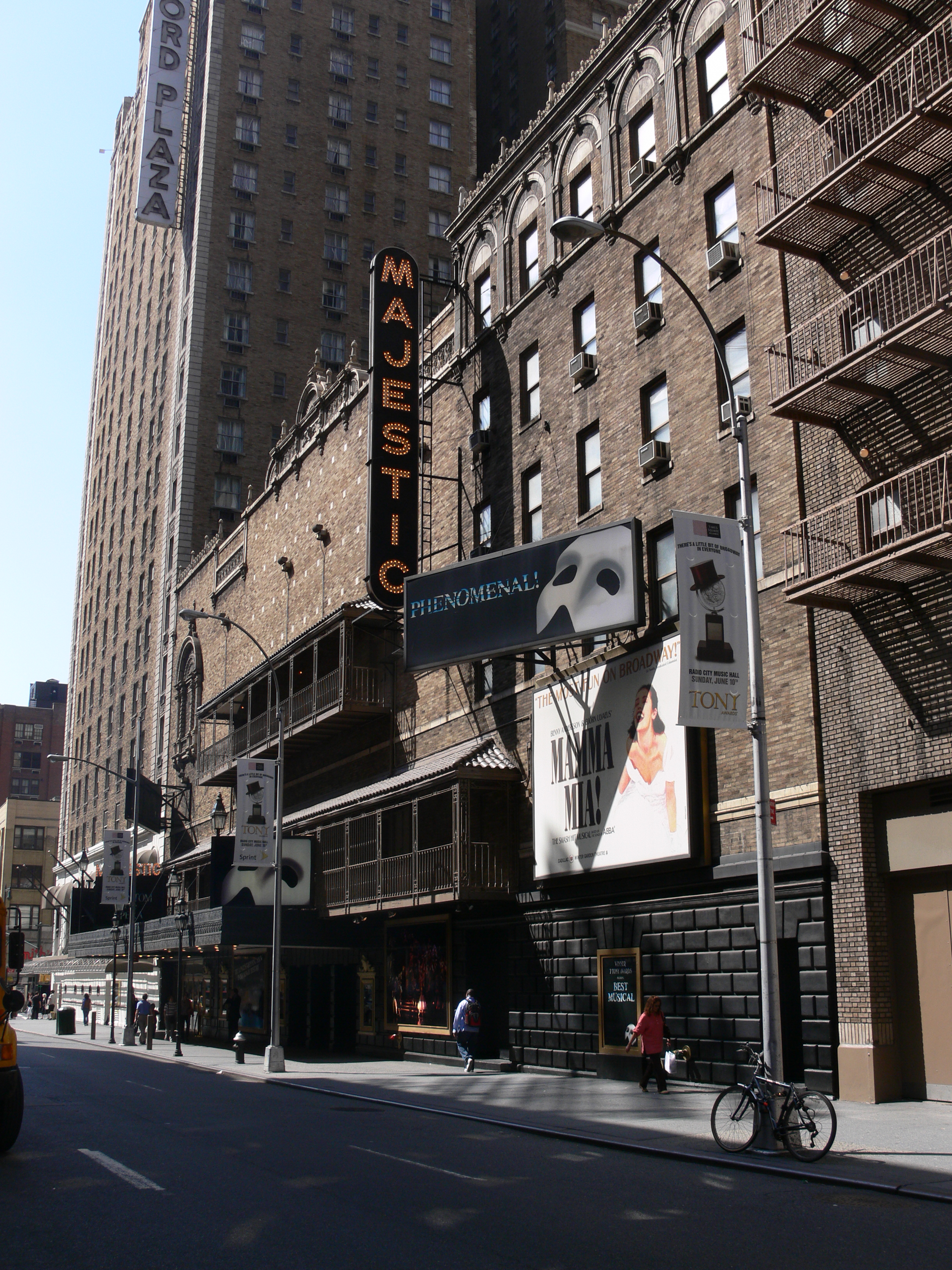The outside of the Majestic Theatre during the daytime on a clear day, with the Row NYC hotel in the background. In addition to various signage and logos for The Phantom of the Opera, a large billboard on the facade includes an advertisement for Mamma Mia! at the Winter Garden Theatre and a banner on a streetlight advertises an upcoming broadcast of the Tony Awards.