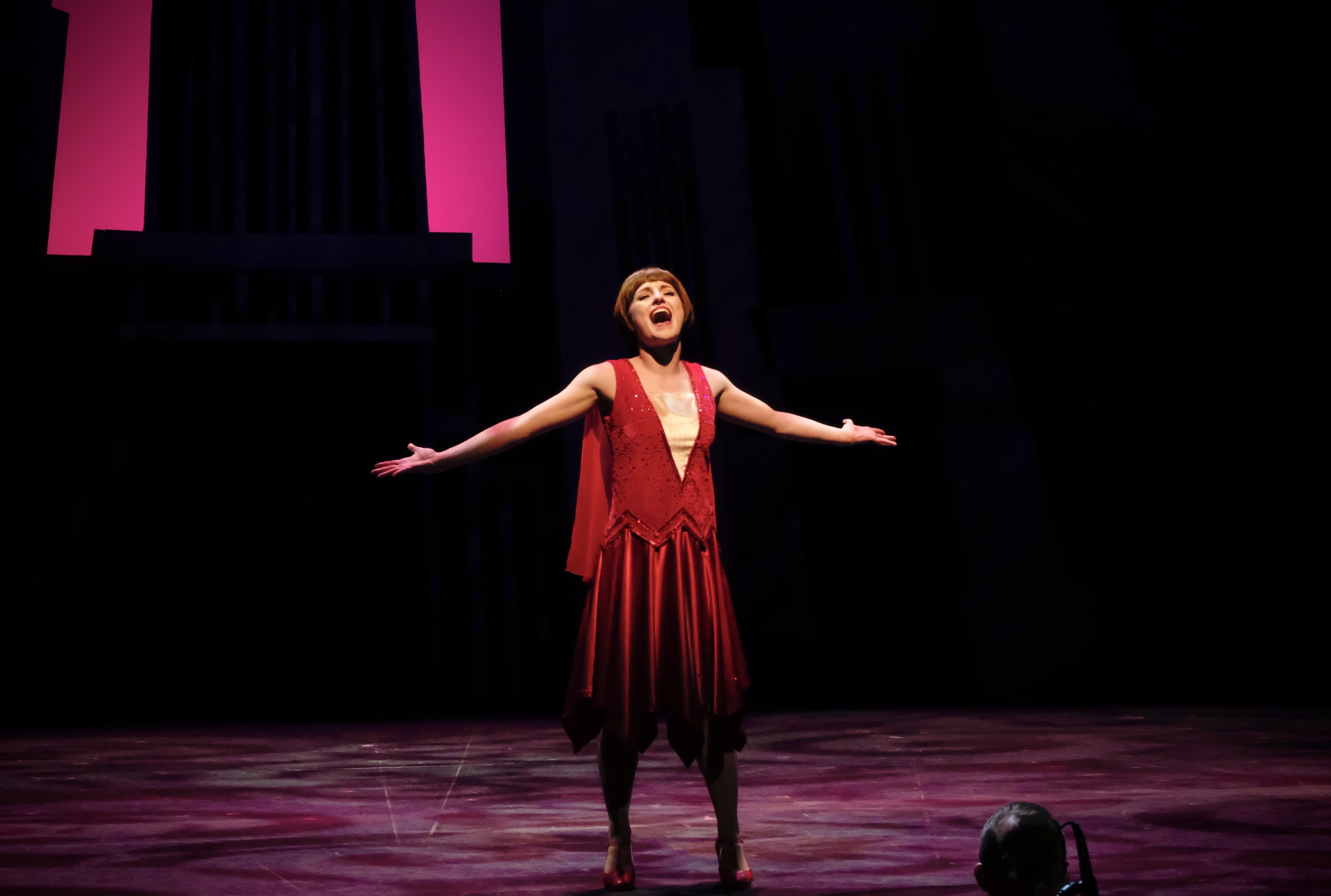 Abigail Isom sings, arms outstretched, on an otherwise empty stage, dressed in a 1920s-era red dress and heels.