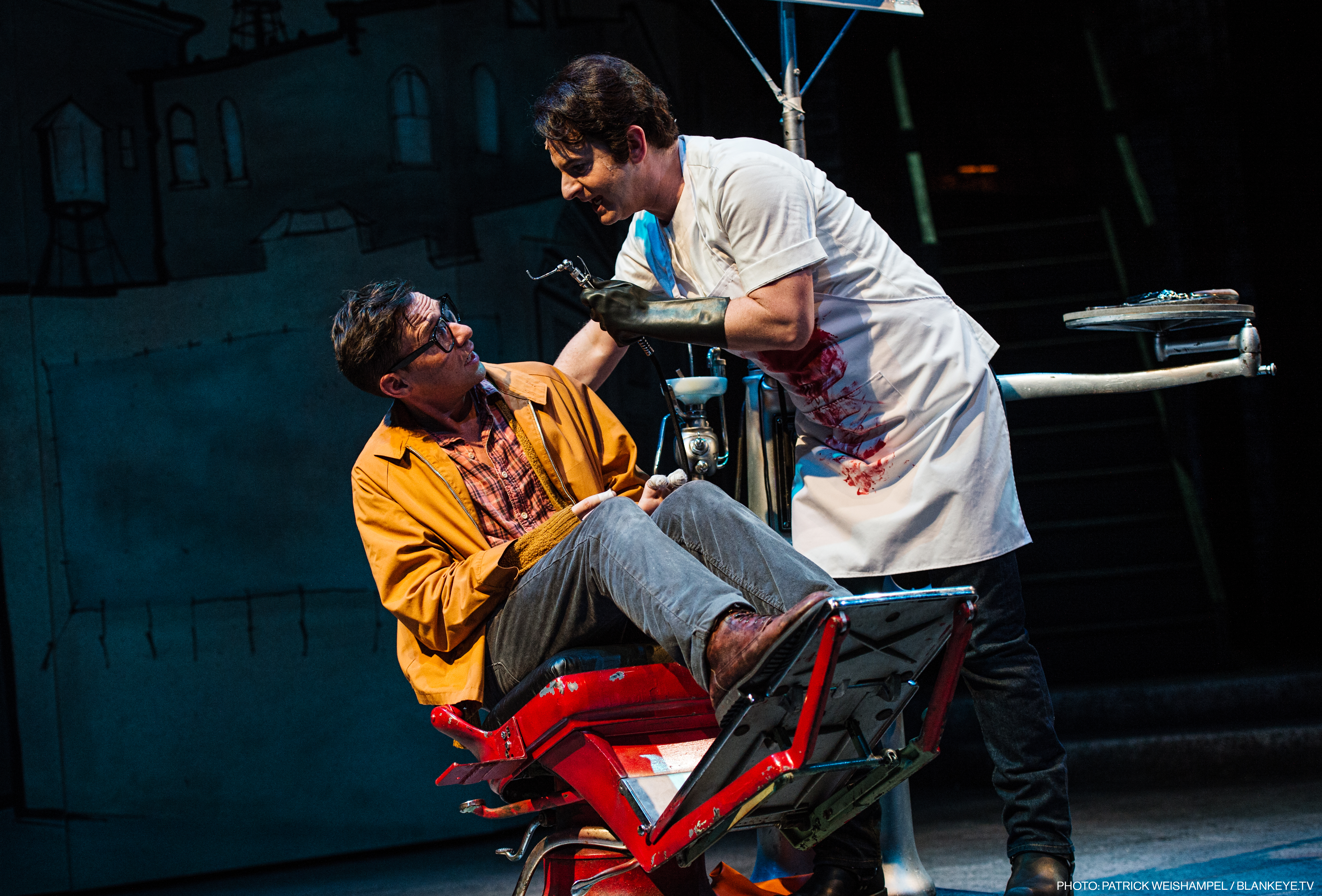 Nick Cearley, portraying Seymour, sits in a dentist chair as Jamison Stern, portraying Orin in a blood-covered apron, looms over him with a frightening-looking dental instrument.
