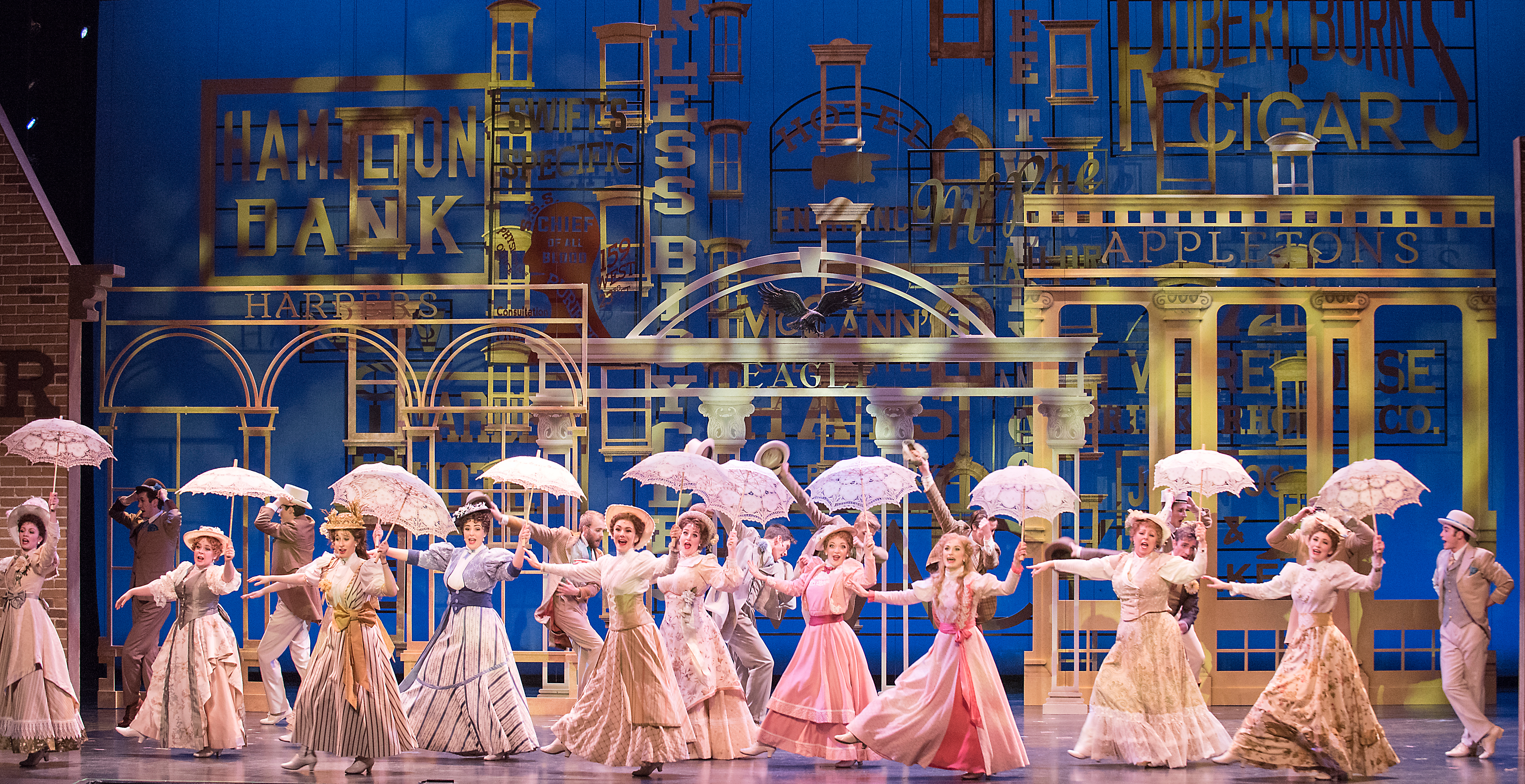 Members of the University of Utah Theatre Department, dressed in late-19th century dresses and carrying parasols, sing a production number in front of a backdrop of period business facades.