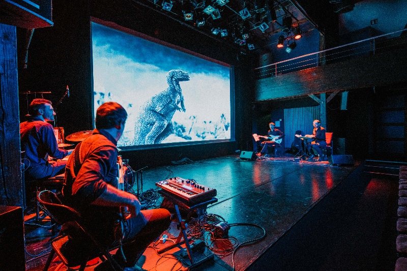 A keyboardist, two guitarists and a drummer perform on stage with a screen displaying a black and white movie scene featuring a large dinosaur.