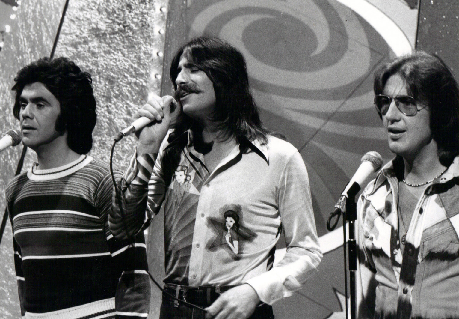 A black and white photograph of Danny Hutton, Chuck Negron and Cory Wells singing into microphones in front of a set featuring patterns and lights.