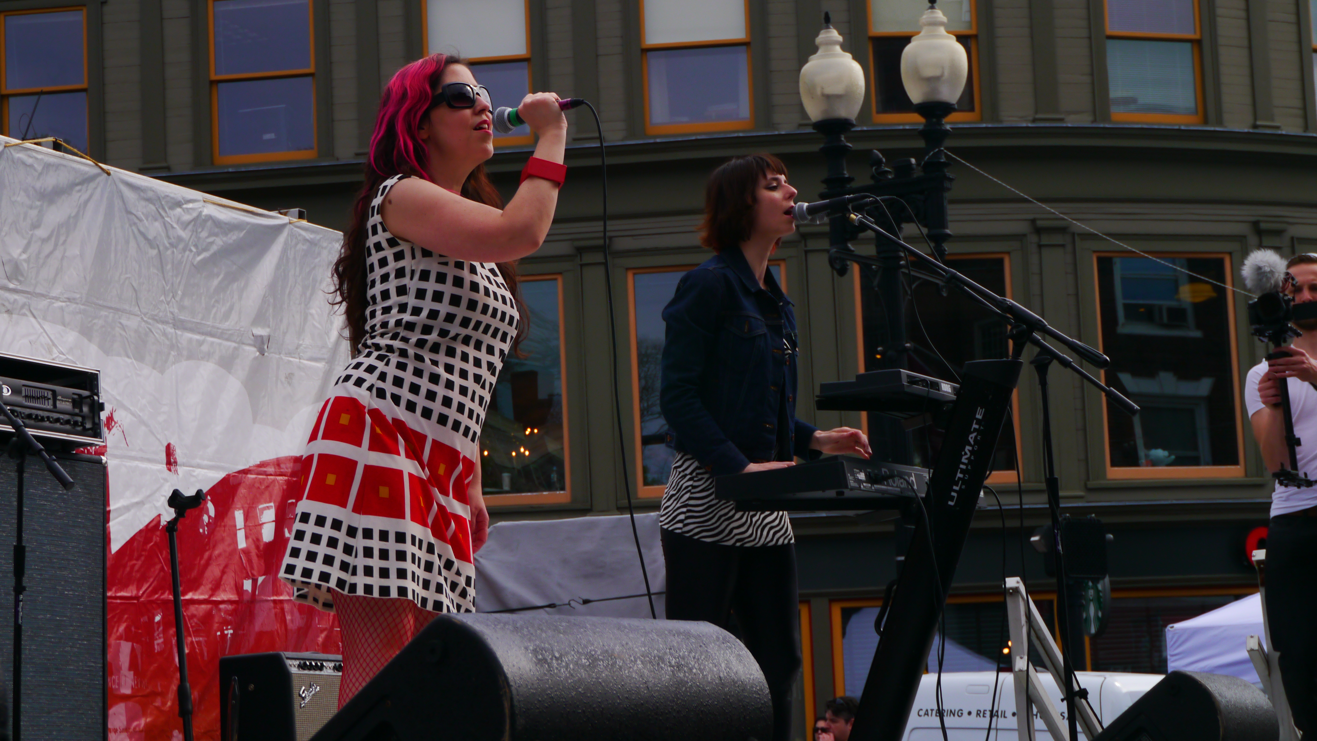 Two members of the band Freezepop sing and play synthesizers on an outdoor stage with a windowed building in the background.