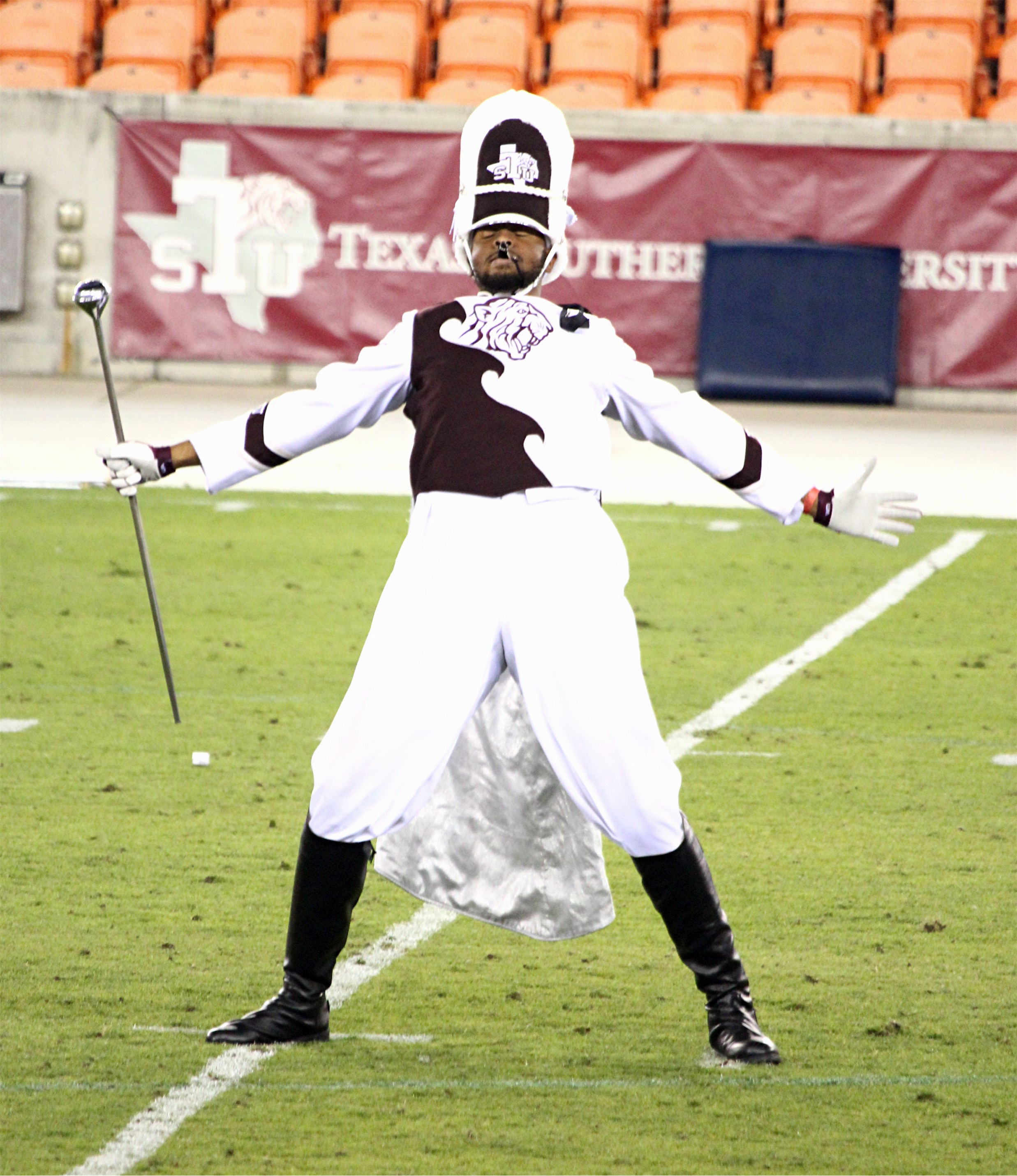 A drum major dressed in a black and white uniform, wearing a busby and holding a mace, standing on a football field, leaning back with his arms outstretched.