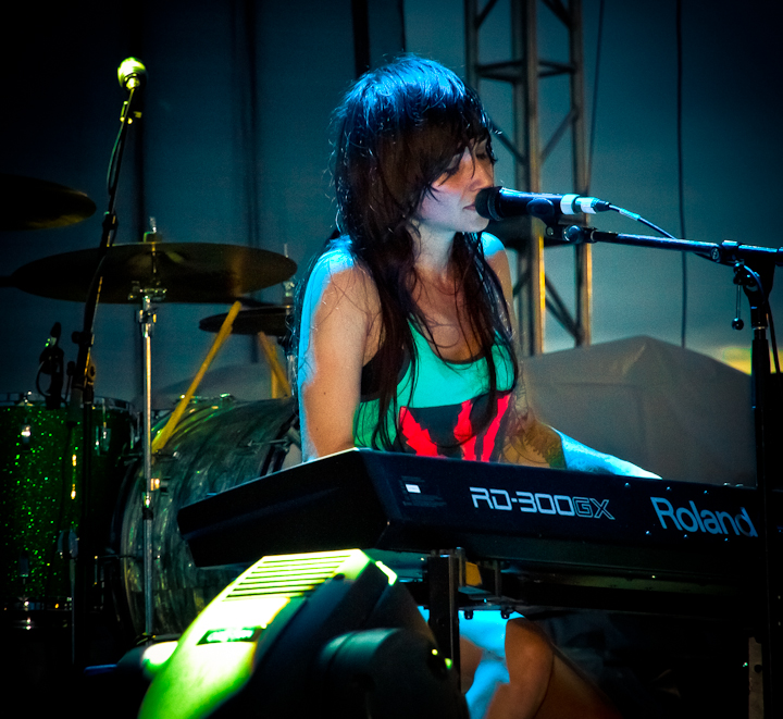 Canadian singer-songwriter Lights sings and plays a Roland RD-300GW keyboard synthesizer on stage. In the background there is a drum set and a metal strut forming part of the stage structure.