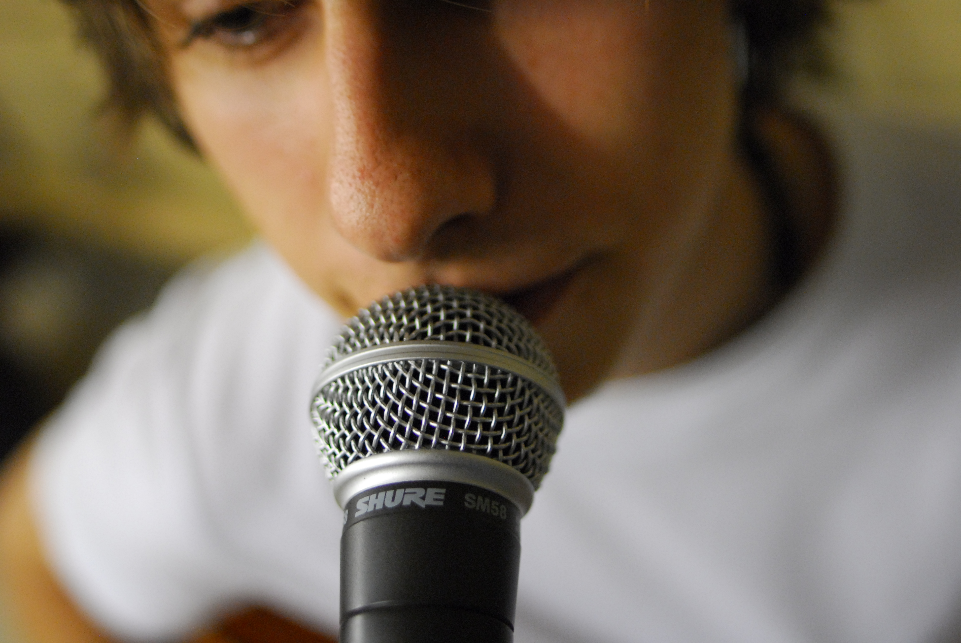 A closeup of a singer singing into a microphone. The photo is focused on the microphone, which has a slate-colored body and a globe covered with a light gray metallic netting. The image of the singer is slightly blurred, and the  microphone's branding, a Shure SM58, is clearly legible at the top of the microphone's body.