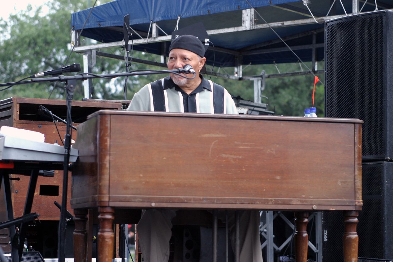 A photograph of Art Neville playing a Hammond organ and singing into a microphone on an outdoor stage. The organ console is flanked on the right by large speakers and on the left by a white synthesizer keyboard. Behind Neville is a taller cabinet with a wood grain finish that matches the Hammond organ.