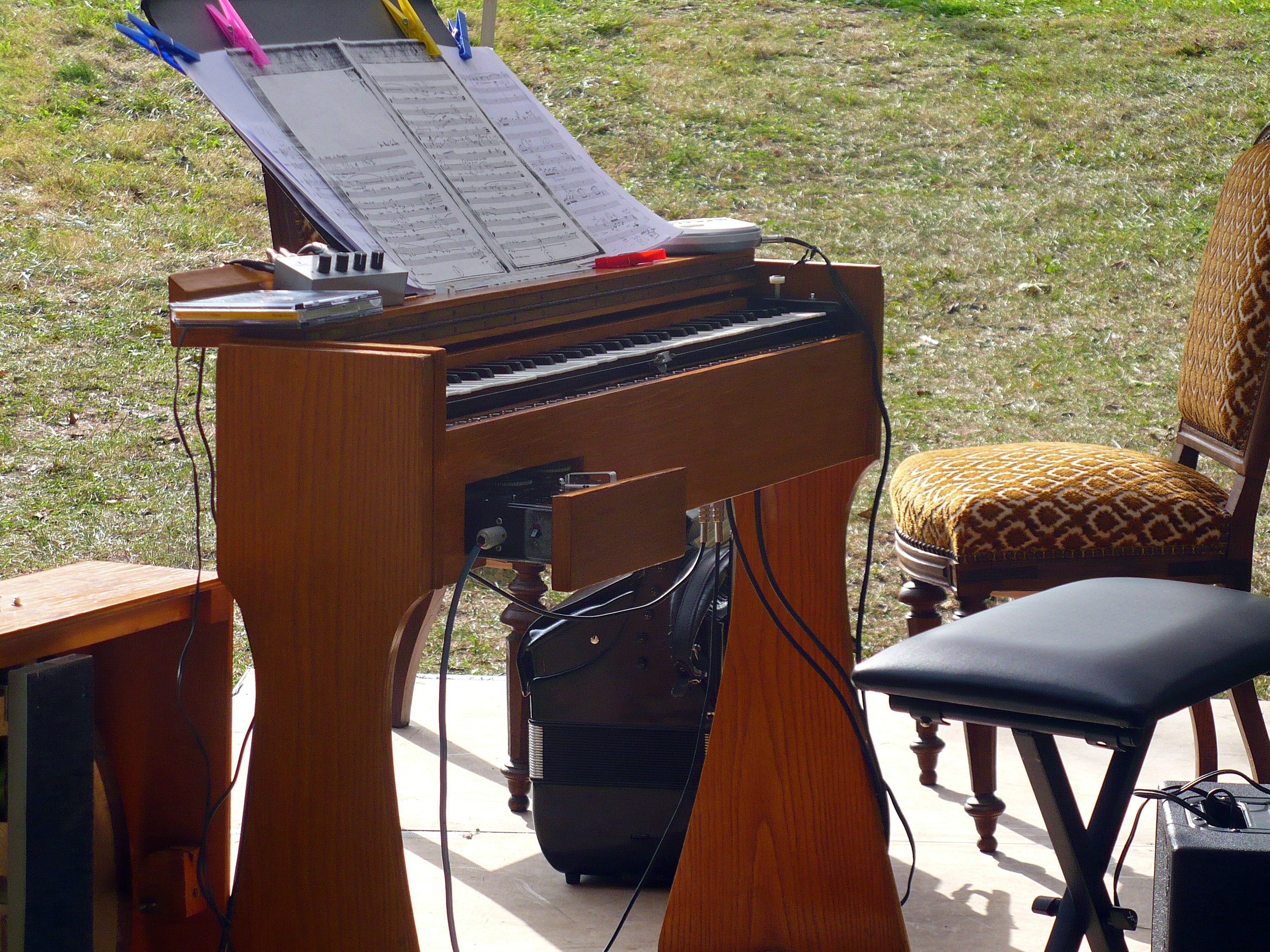 A photograph of the keyboard console of an ondes Martenot sitting on an stage. No one is seated at the console, but photocopied pages of sheet music sit on the stand, held in place by colorful clothespins. The rear of the ondes' speaker is seen in front of the console.