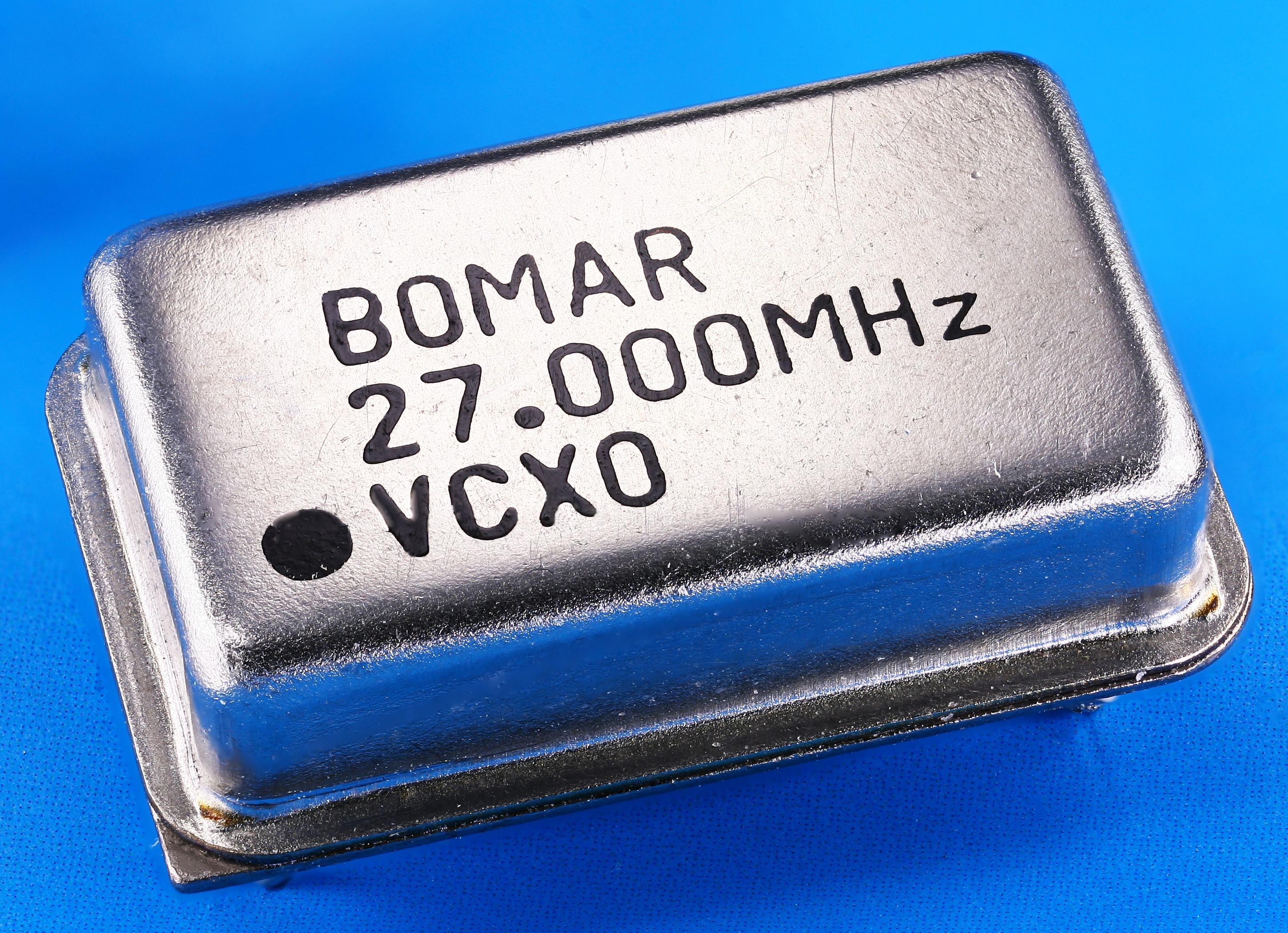 A close-up photograph of an oscillator component. Seen from above, the component is a shiny metal unit with a top cover that looks like an up-turned miniature cakepan. On the top of the component is the label `BOMAR 27.000MHz VCXO`.