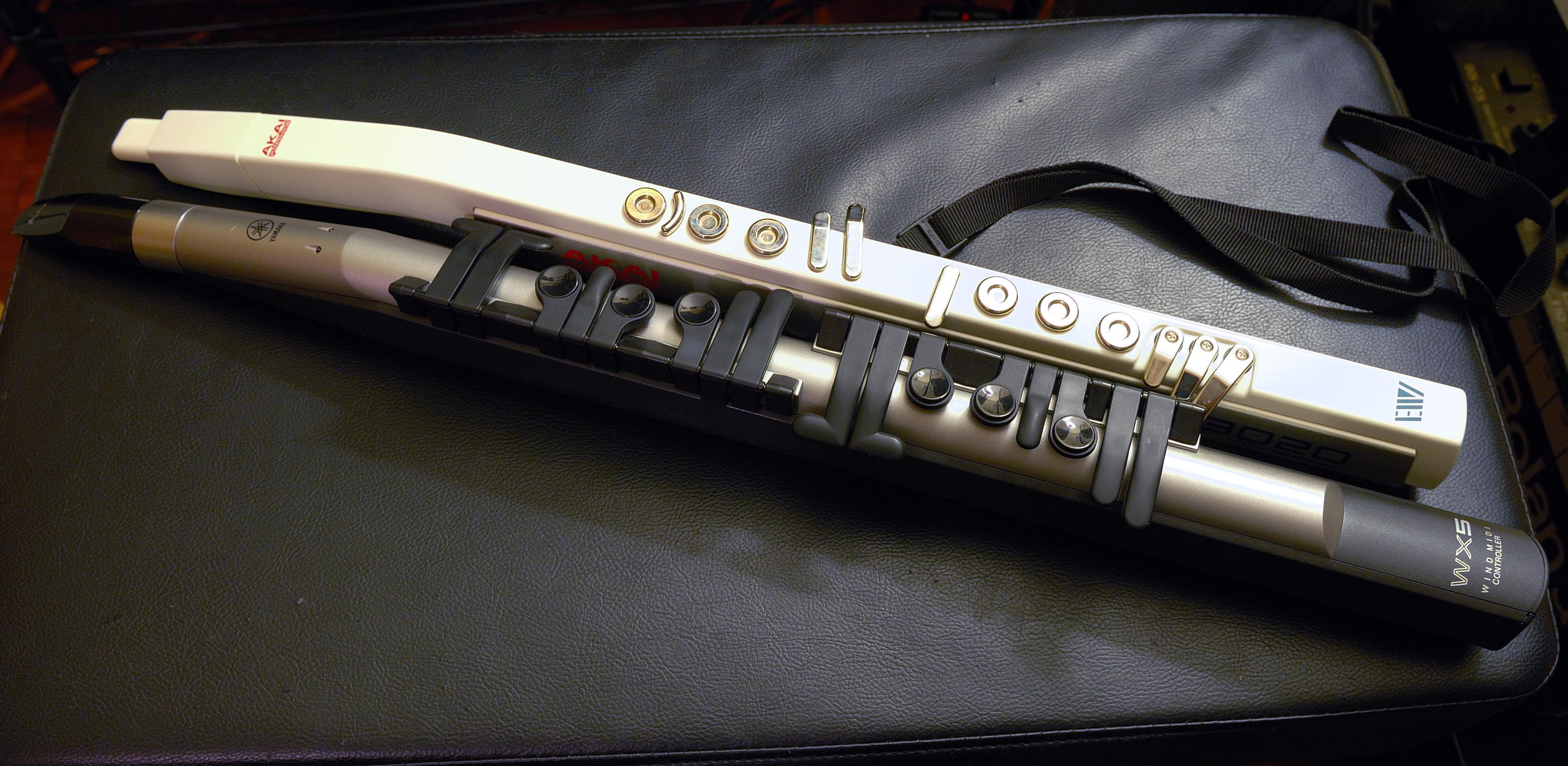 A photograph of two wind controllers, an Akai EWI 3020 and a Yamaha WX5, laying on a leather cushion. Both have plastic bodies; the Yamaha instrument has sixteen visible keys in a lever configuration while the Akai instrument has thirteen touch-senstitive metal discs.