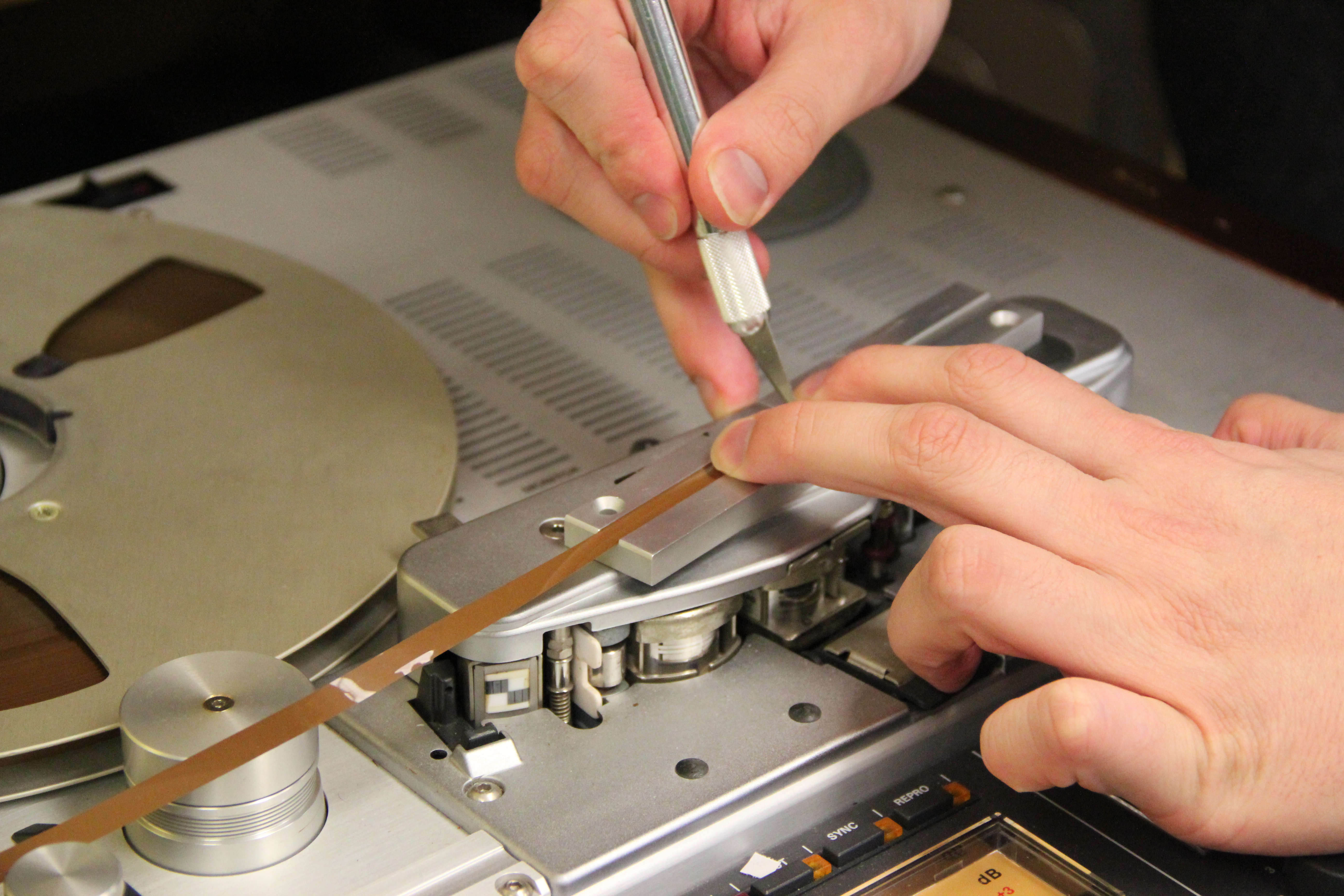 A close-up of a reel-to-reel tape player laying on it's back. A full supply reel is in place on the left-hand spindle of the tape recorder, and one end of the tape is being held by someone left hand in a splicing block while being cut by a hobby knife held in their right hand.