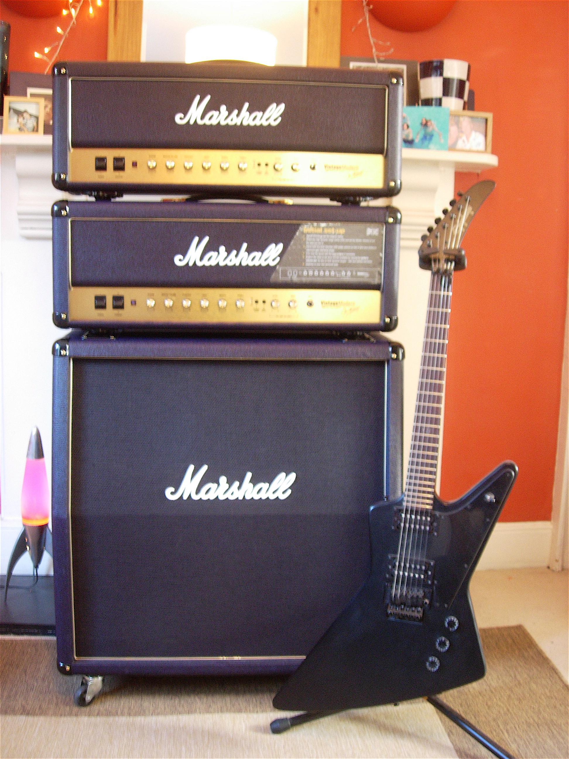 A black electric guitar on a guitar stand, sitting in front of a stack of audio equipment: from top to bottom, a Marshall Vintage Modern 50 watt amplifier, a Marshall Vintage Modern 100 watt amplifier, and a Marshall cabinet loudspeaker.