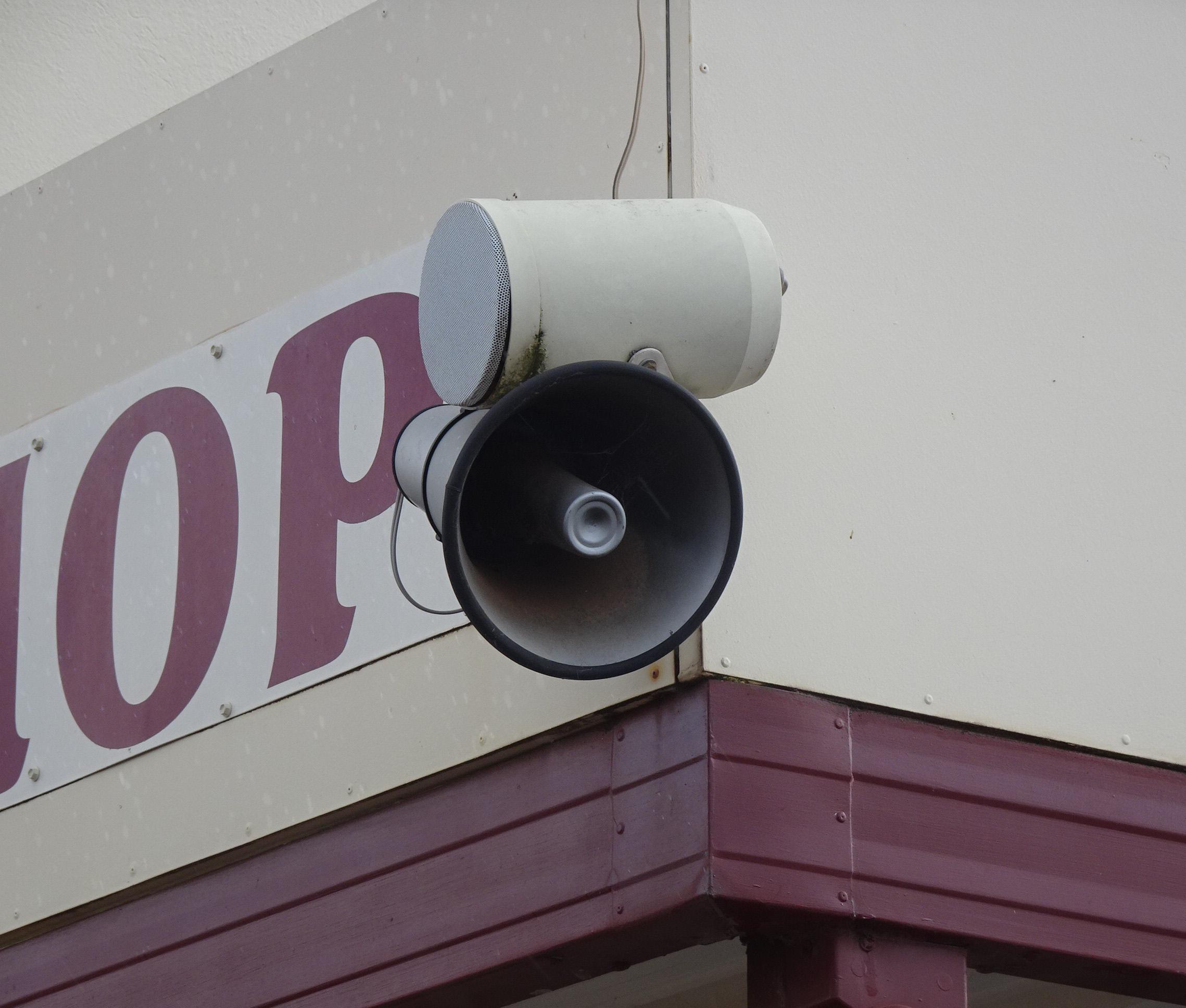 Two speakers, one cylindrical with a mesh cover, and another in the shape of a megaphone, mounted to the corner of a building.