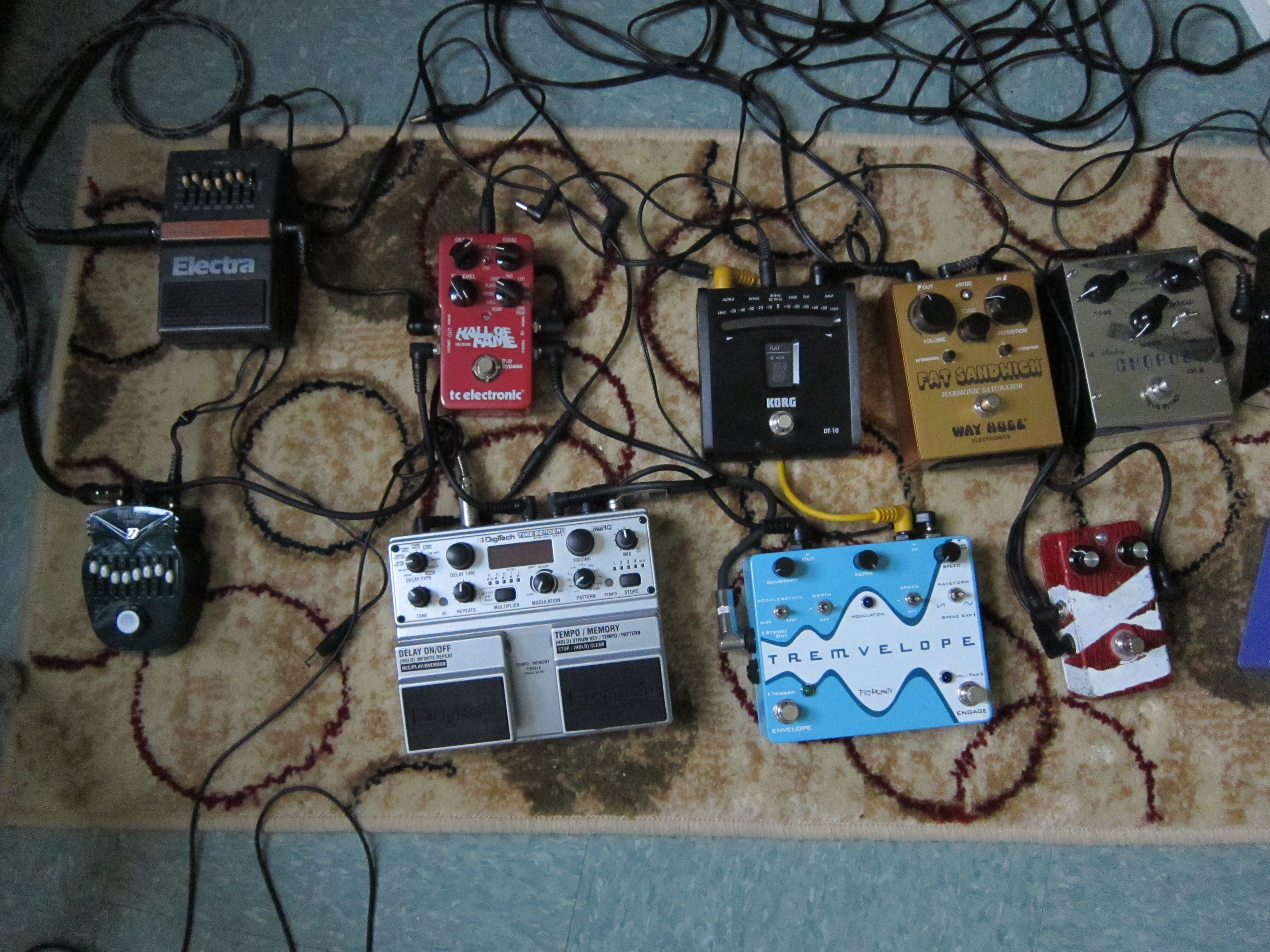 A collection of nine different guitar pedals amidst a collection of various cables, some connected to pedals and other loose, on a small patterned rug. The pedals vary in size and color and have an assortment of controls: small dials, knobs and sliders meant to be controlled by hand, and large buttons intended to be pressed by the feet while playing.