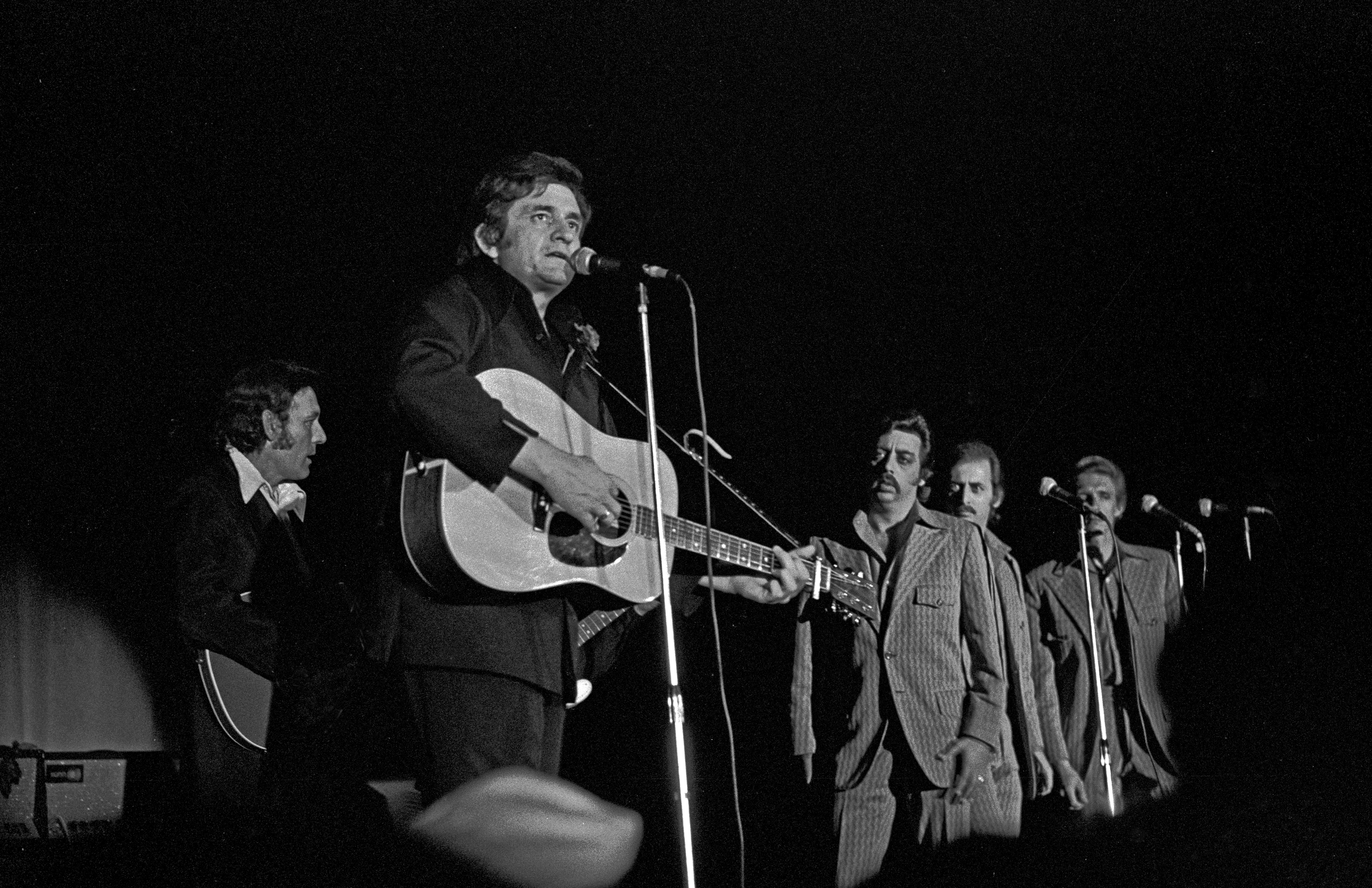A black and white photograph of Johnny Cash seated on a stool playing an acoustic guitar and singing into a microphone. Behind cash, Carl Perkins plays guitar, and Harold Reid, Don Reid, and Philip Balsley stand watching Cash perform.