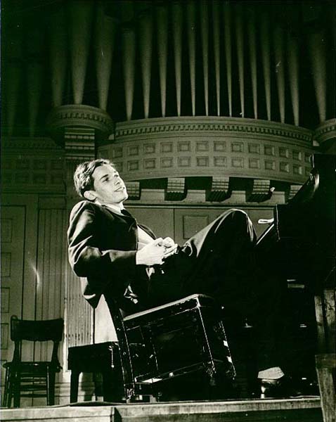 Pianist Glenn Gould smiles and leans back in his chair at a grand piano, on stage with ranks of organ pipes in the background.