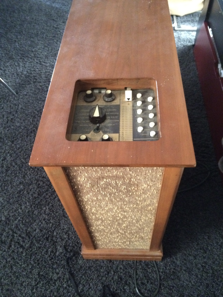 A Wurlitzer Side Man unit: a wooden enclosure about three feet high, three feet deep and 18 inches wide, with an inset control panel on the top with several buttons and dials.