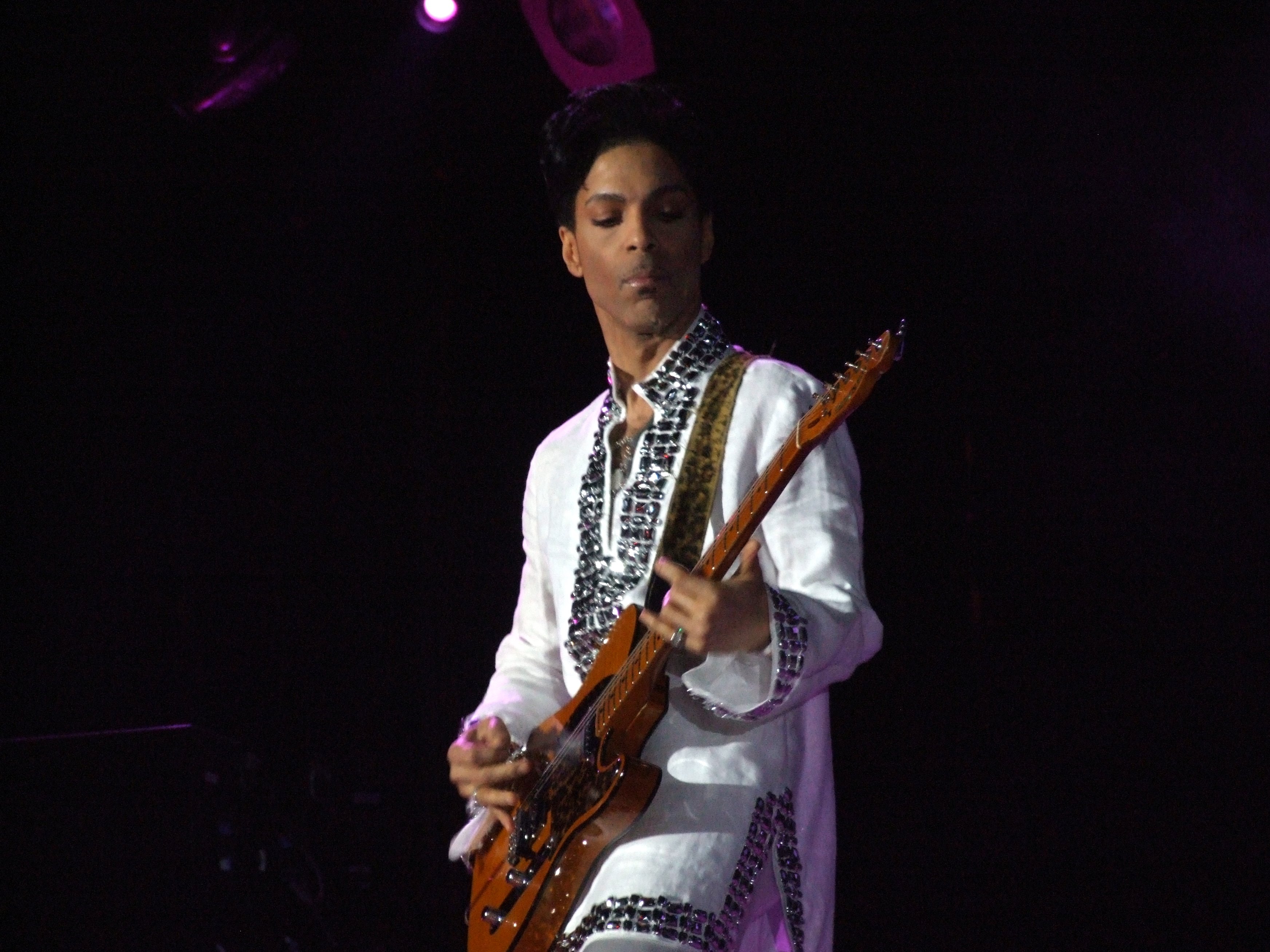 Prince, playing guitar against a dark background, wearing a white shirt bordered with large gems.