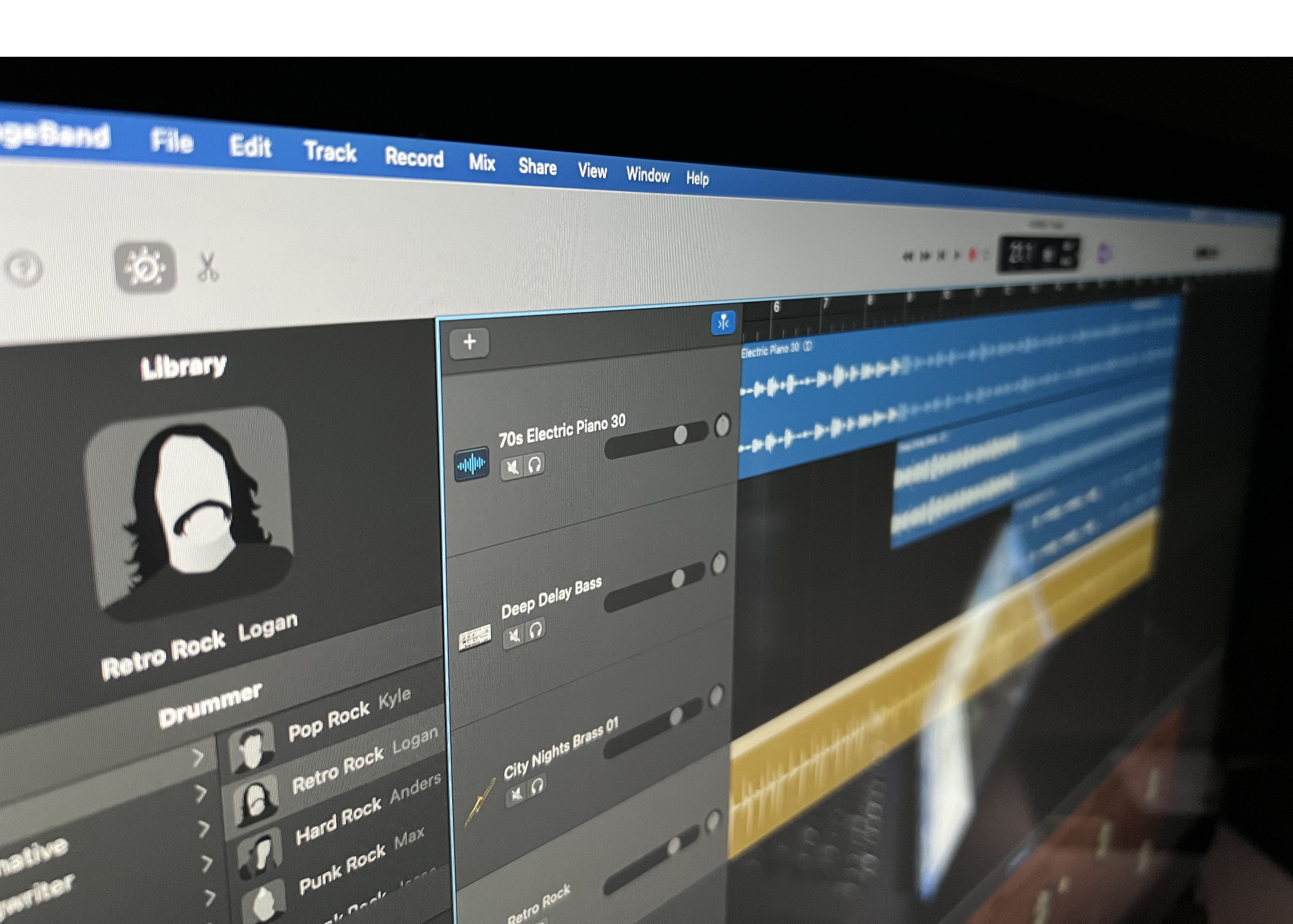 A close-up photograph of a display showing the GarageBand interface. GarageBand is showing a project with four tracks, labelled `70s Electric Piano 30,` `Deep Delay Bass,` `City Nights Brass 01,` and `Retro Rock.` The latter is selected, and the right side of the screen lists displays the virtual drummer as `Logan,` who is pictured as a silhouette of a man with long, wavy hair and a handlebar mustache.