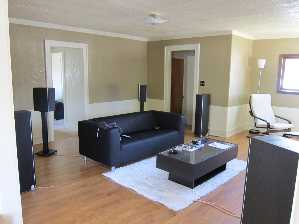 A photograph of the seating area of a home theater setup. A black leather sofa sits in the middle of the room with a coffee table in front of it on a white shag area rug. Floor speakers are placed directly to the right and left of the sofa, and two shorter speakers on stands are placed at the two rear corners of the sofa. The corner of a front speaker is visible in the bottom right corner of the photograph.