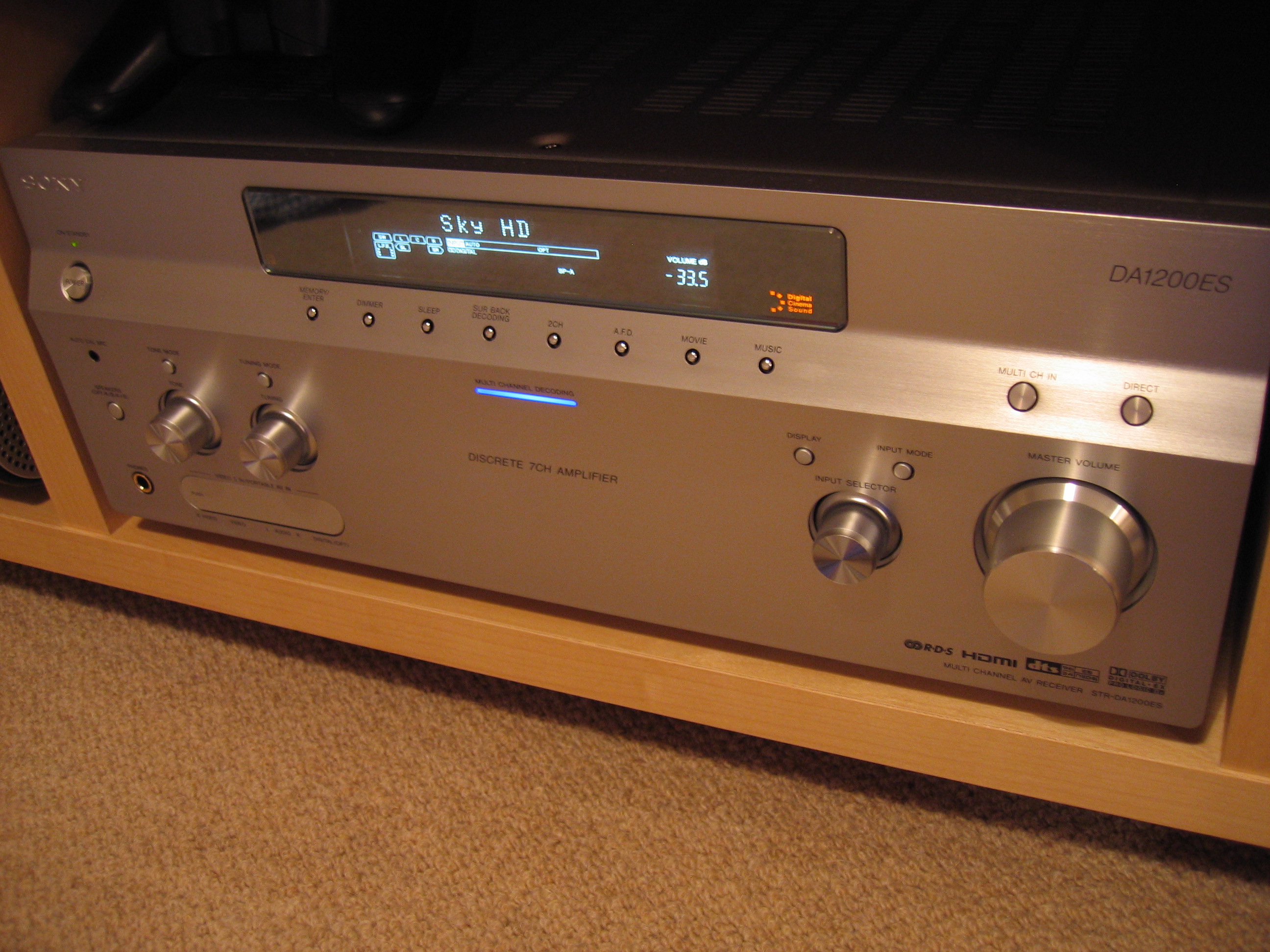 A silver audio component in a home theater system, 18 inches wide, 18 inches deep and 6 inches tall. The front of the unit has four dials, sixteen buttons, and an LED display. Below the display, a blue light labeled 'multi channel decoding' is glowing.
