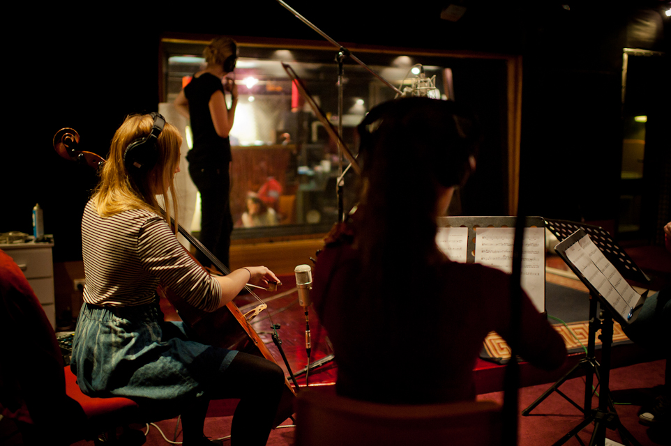 A photograph of a cellist and violinist, both wearing headphones, performing in a recording studio. The musicians are seated facing away from the camera, and a large window into another room is visible in the background.