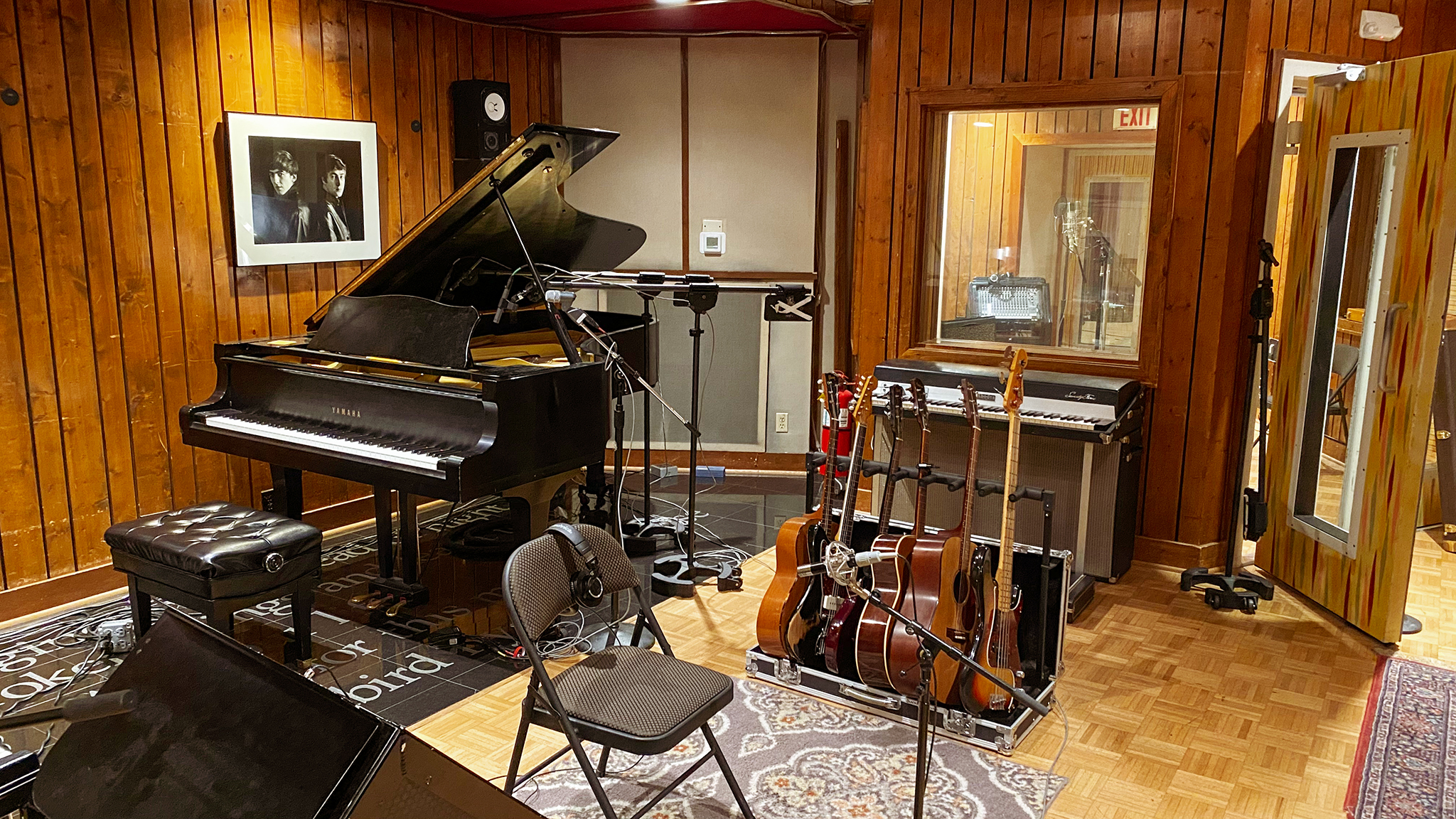 A room with wood floors and wood paneling containing a baby grand piano with microphones for the piano strings and a seated vocalist, a guitar stand holding three acoustic guitars, two electric guitars and an electric bass, and an electric piano. Adjacent to the room is a vocal isolation booth with an open door.