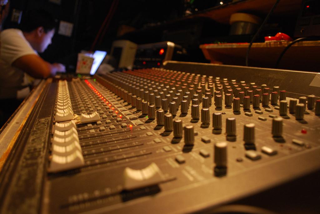 A close-up photograph of a large 20-channel mixing board with a recording engineer at a computer station in the background.