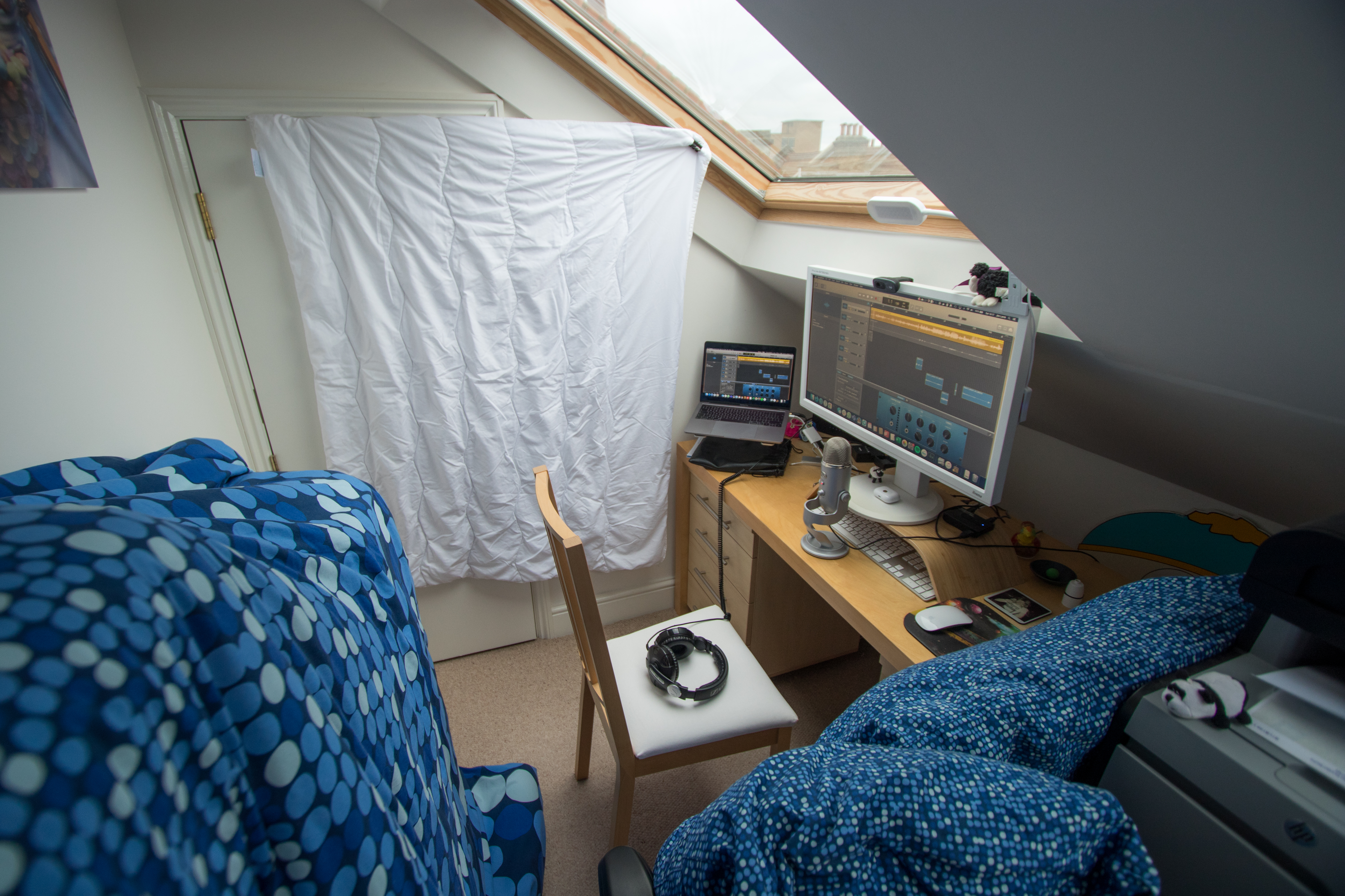 An attic room with a slanted ceiling and large outside window containing a computer station with a laptop, monitors, and microphone. The wall beside the computer station and the furniture surround it are covered with various comforters and pillows.