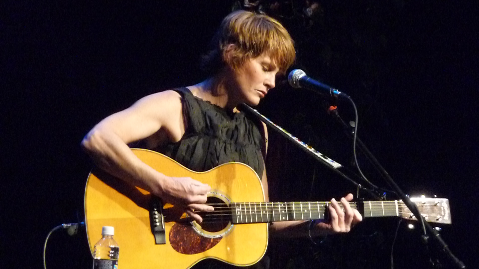 Shawn Colvin plays an acoustic guitar on stage against a black background.