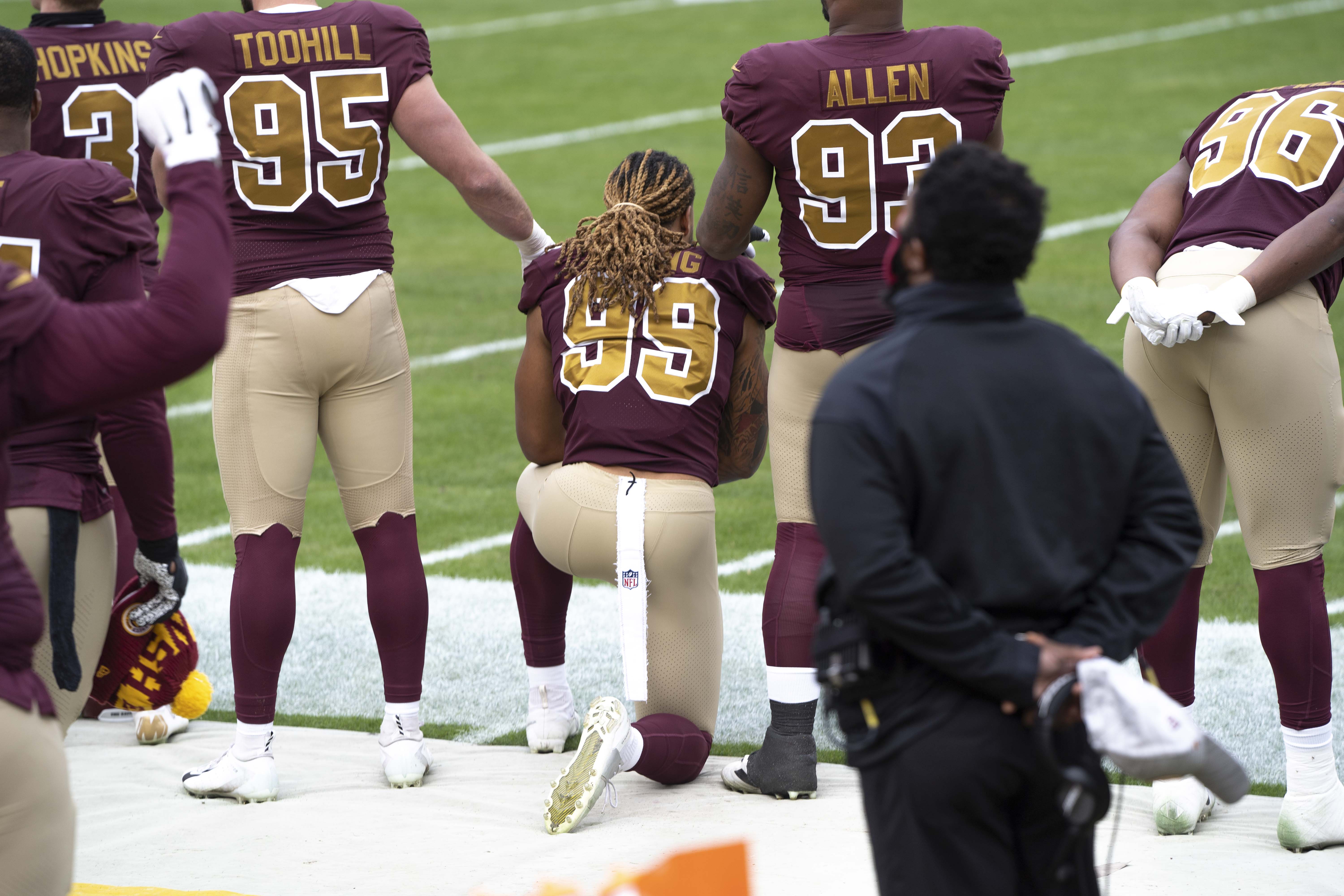A photograph of from behind of football players Dustin Hopkins, Casey Toohill, Chase Young, Jonathan Allen, and James Smith-Williams lined up on the side of the field for the national anthem. Young is kneeling while others are standing, with Toohill and Allen placing their hands on Young's shoulders.