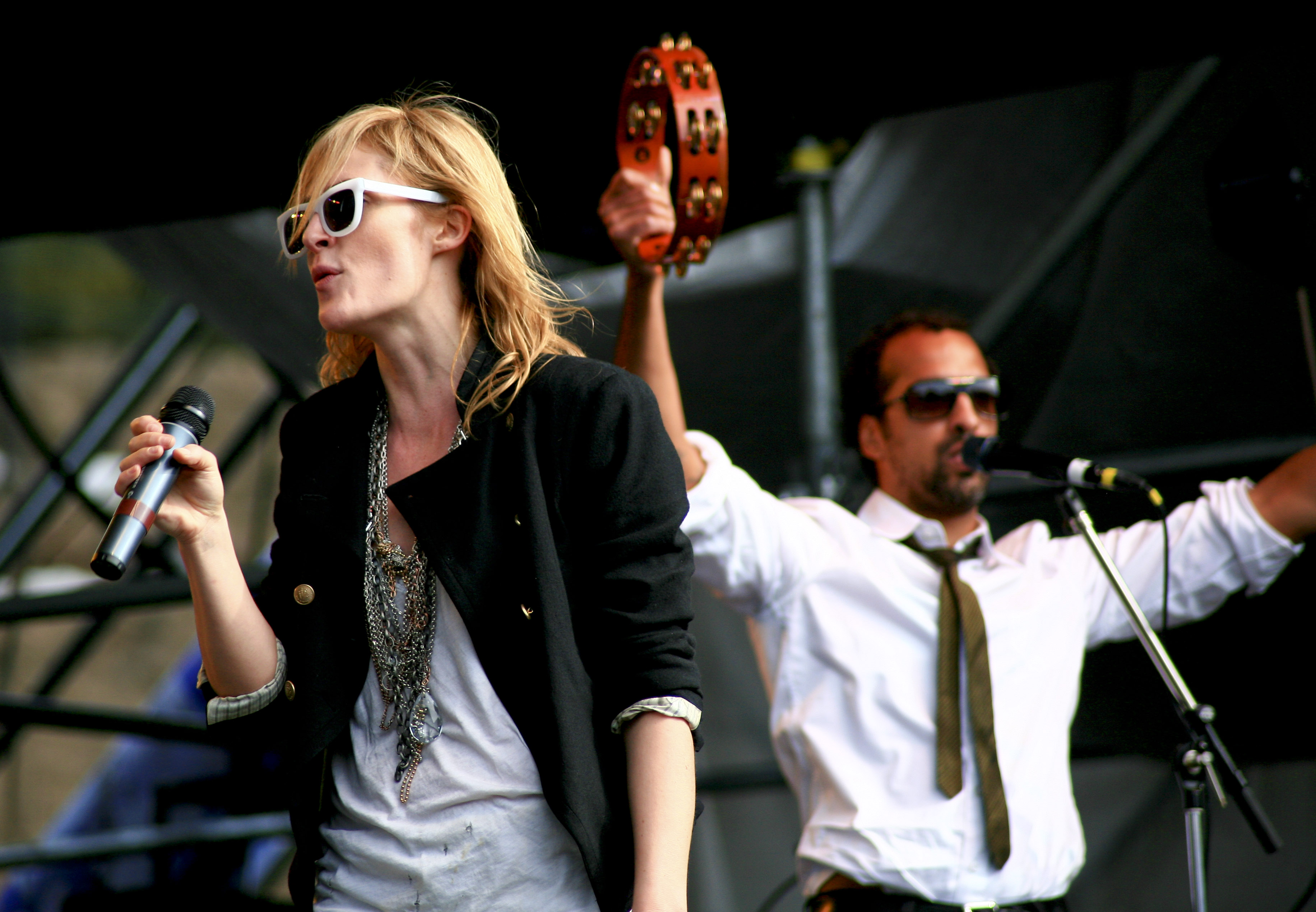 A photograph of Emily Haines and Joshua Winstead performing on stage. Both are singing into microphones, and Winstead is playing a tambourine.