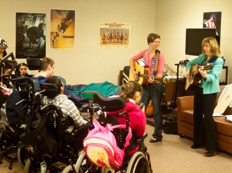 A photograph of two women performing with acoustic guitars before a group of six young people seated in wheelchairs.