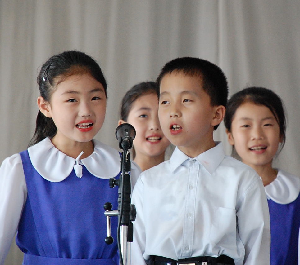 A photograph of three girls and a boy, who appear to around ten years old, singing into a single microphone in front of a gray curtain.
