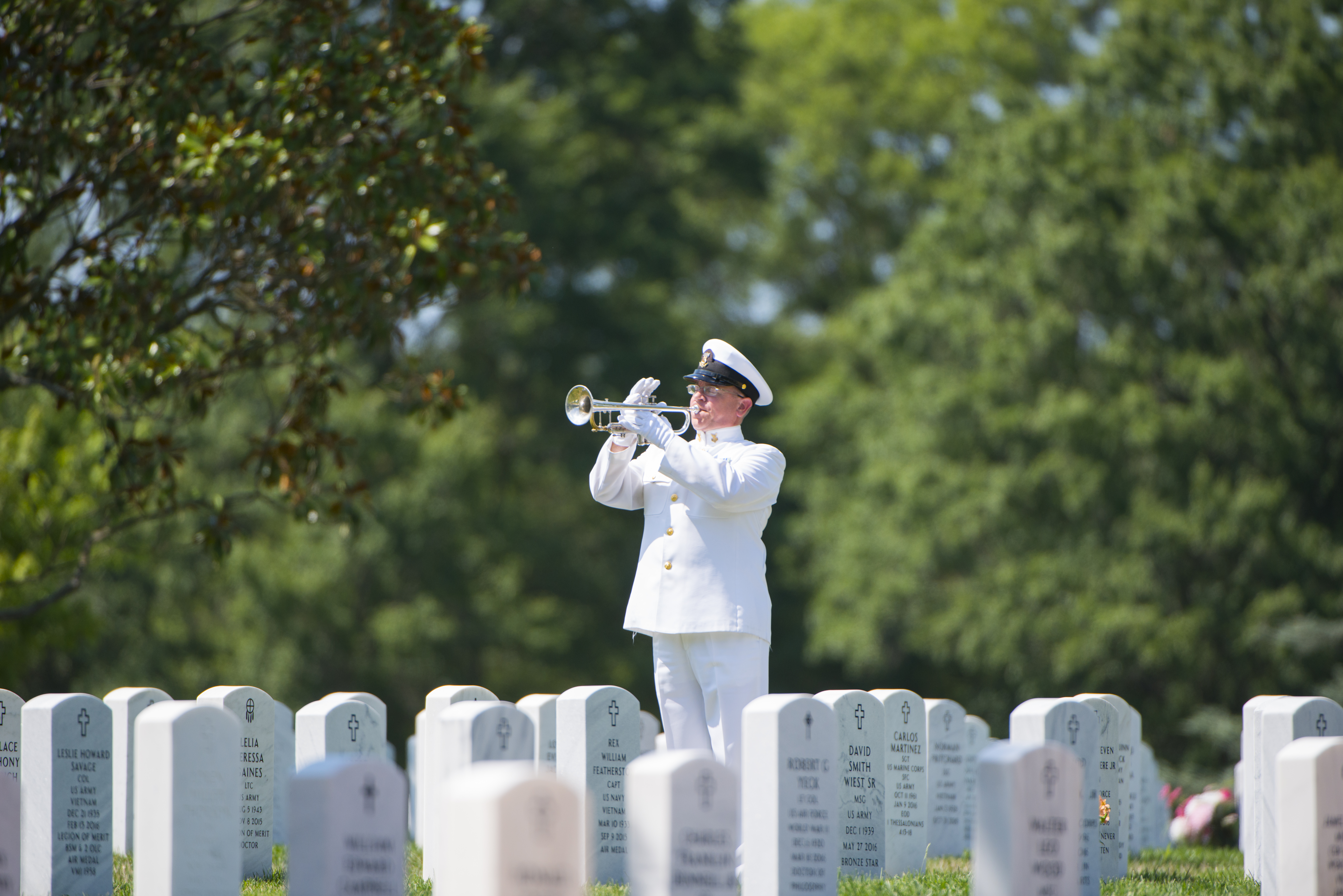 A photograph of a man in a white uniform standing in a military cemetery, playing a silver trumpet.