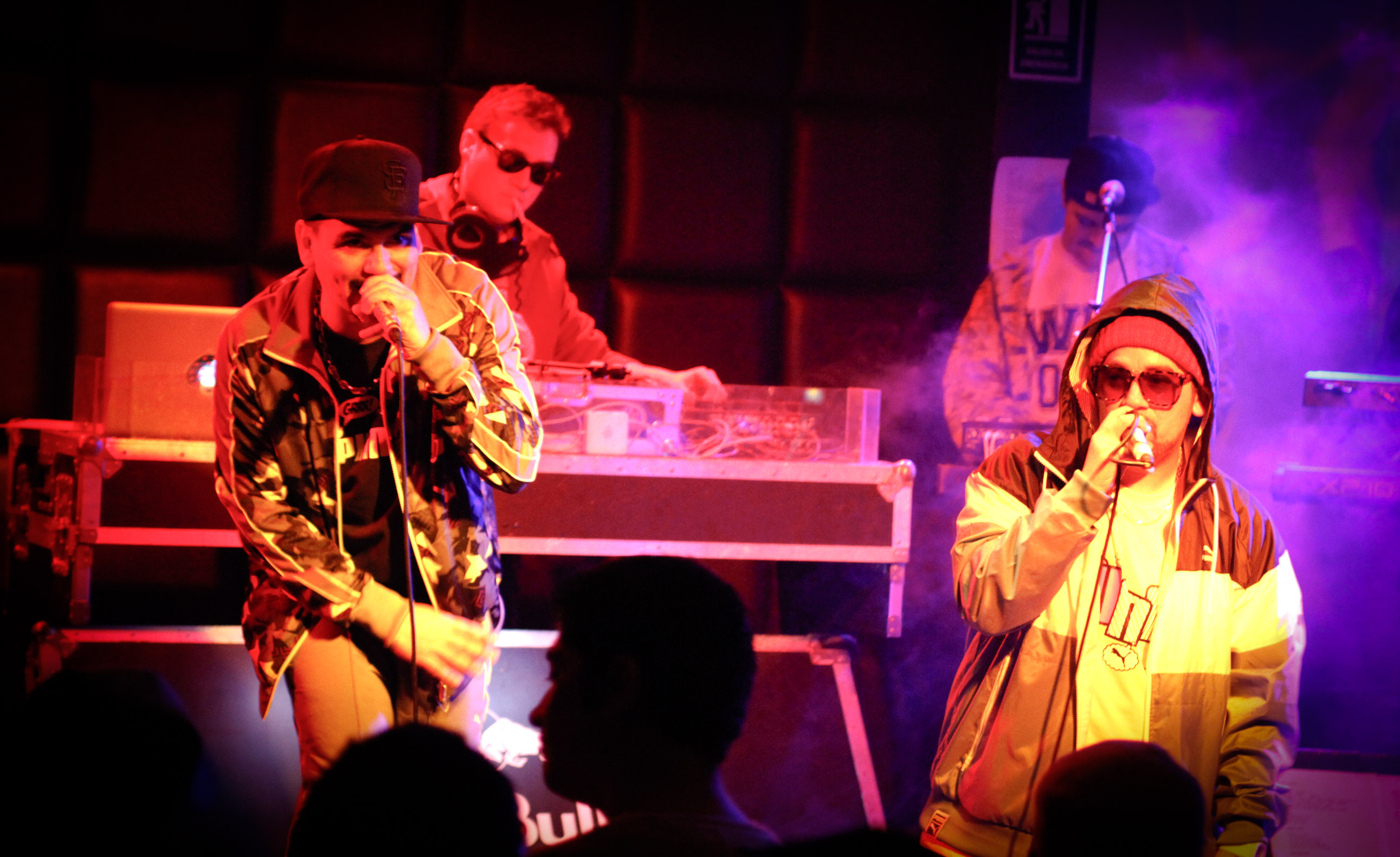 A photograph of Seo2 and MC Nauck rapping into microphones on stage while DJ Caso and Nick Calaveras perform on separate DJ rigs in the background.
