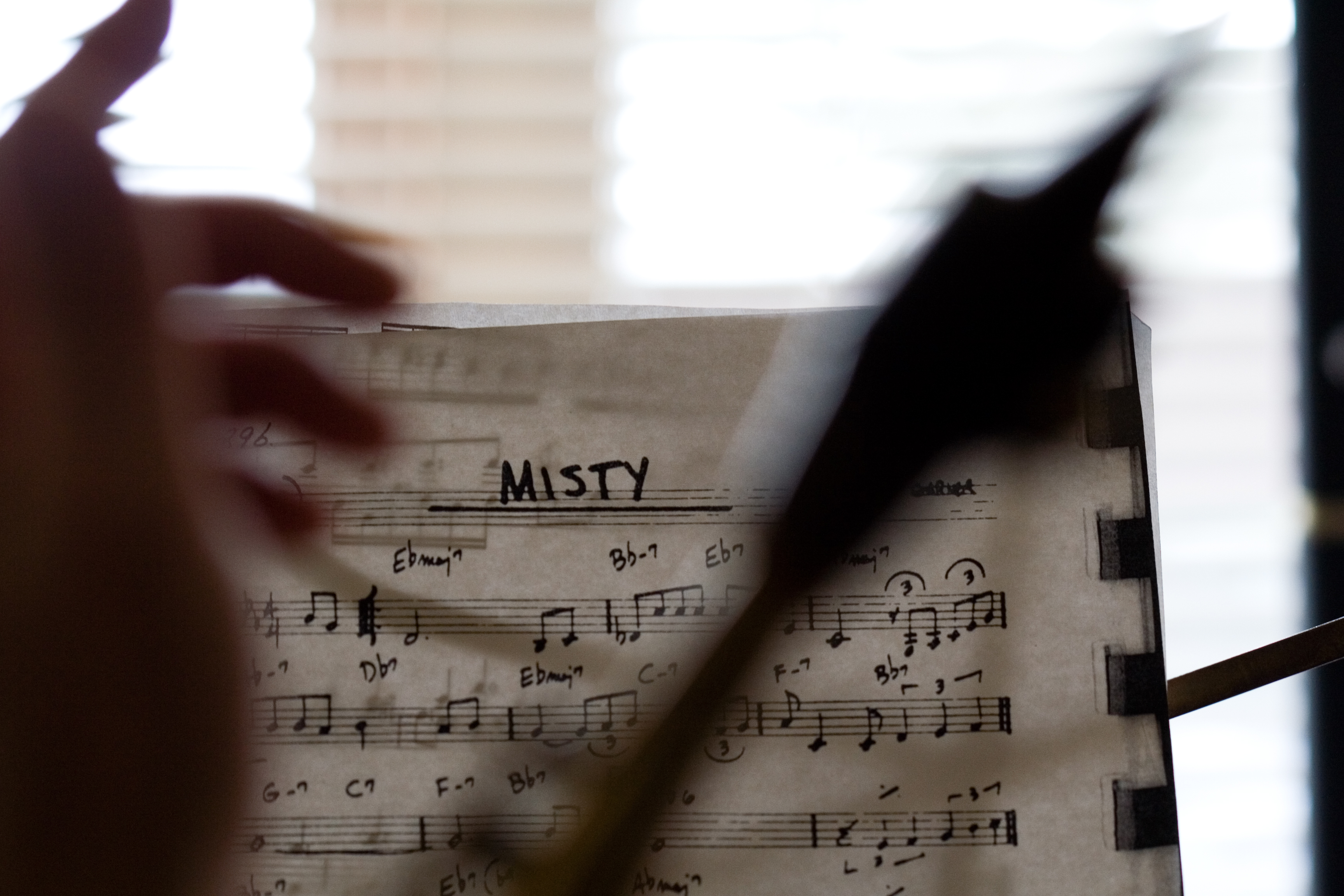A photograph of the sheet music for the jazz chart `Misty` on a music stand, with the blurry shapes of a hand and the neck and mouthpiece of an alto saxophone in the extreme foreground.