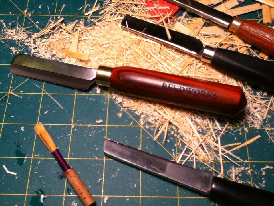 A photograph of an oboe reed laying next to four reed knives and a large amount of cane savings on a green cutting board with yellow grid markings.