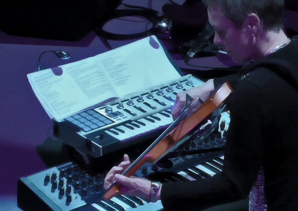 A photograph of Laurie Anderson from behind and to her left as she performs on violin while standing behind two synthesizer keyboards.