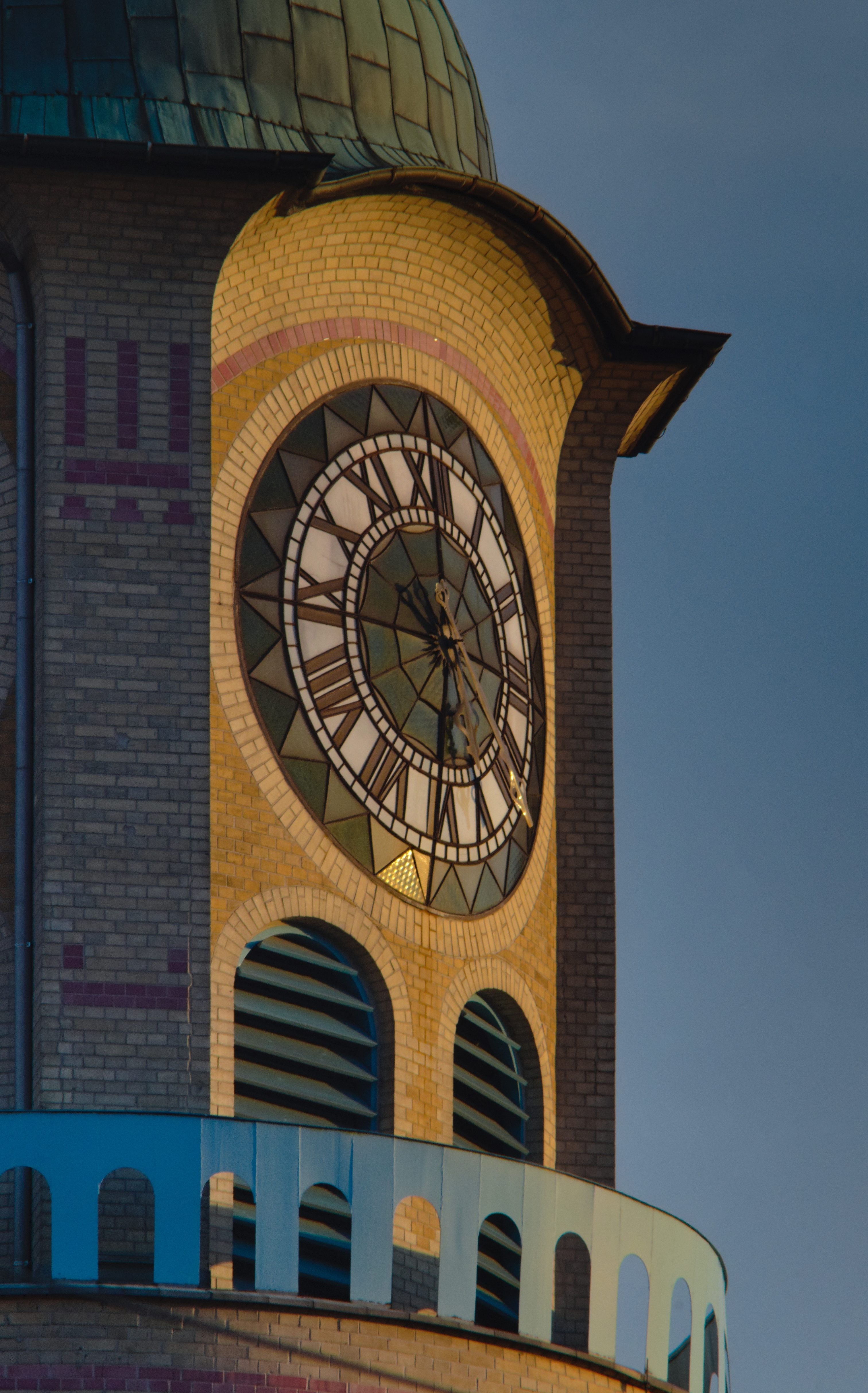 A photograph of a clock tower made of ornate, colorful brickwork and featuring a clock face made entirely of stained glass.