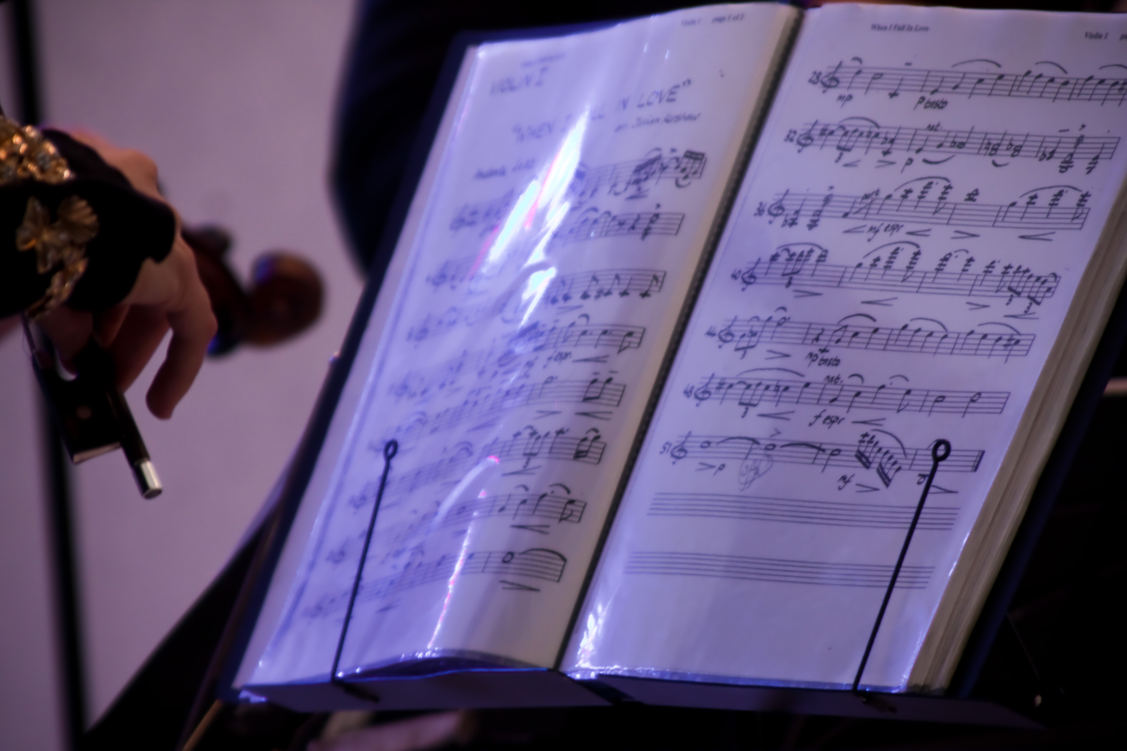 A photograph of an book of music on a music stand open to a page with the first violin part for `When I Fall in Love` by Edward Heyman, with the hand of the violinist, the frog of the bow and the scroll of the violin visible on the left side of the frame.