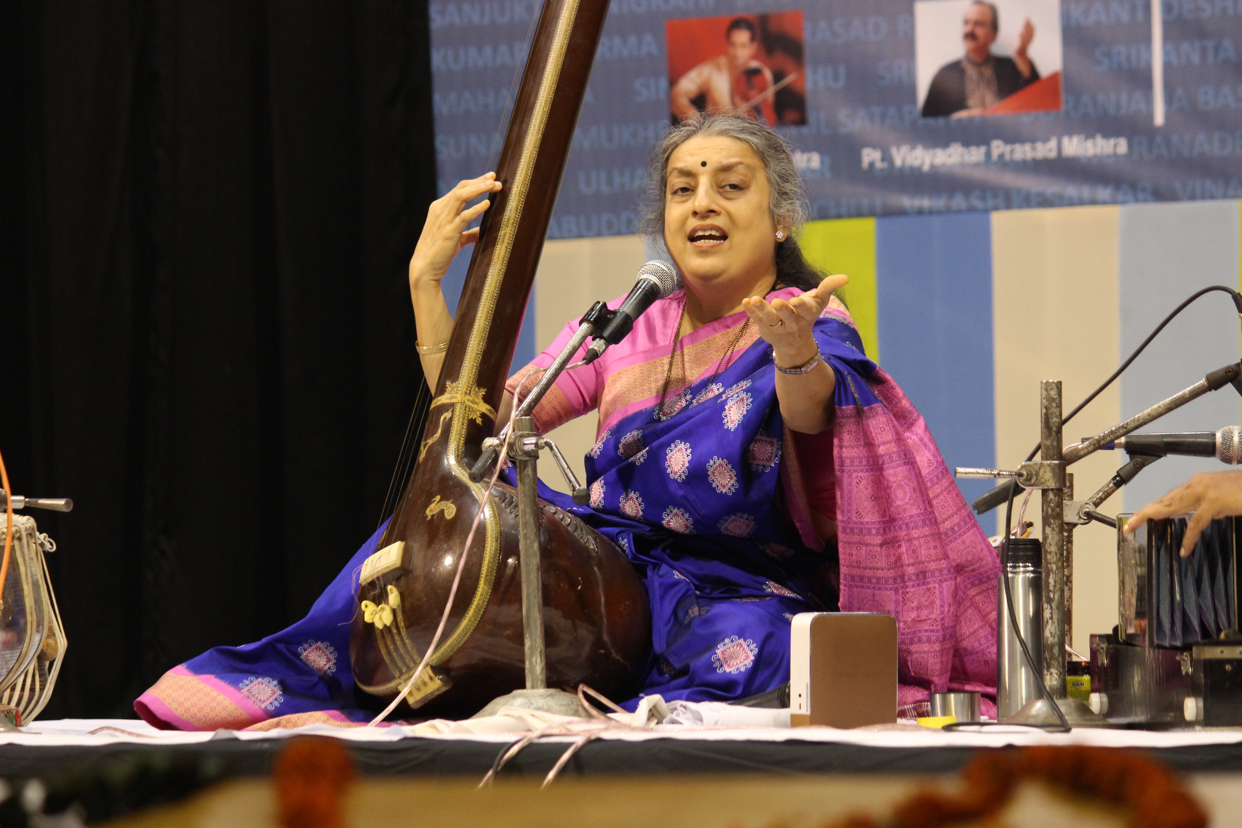 A photograph of Ashwini Bhide-Deshpande sitting on a stage, playing a sitar and singing into a microphone.