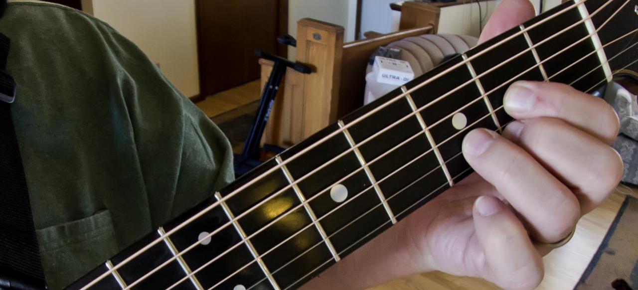 A close-up photograph of a guitarist's left hand with the index finger on the second fret of the G string, the middle finger on the second fret of the high E string, and the fourth finger on the third fret of the B string.